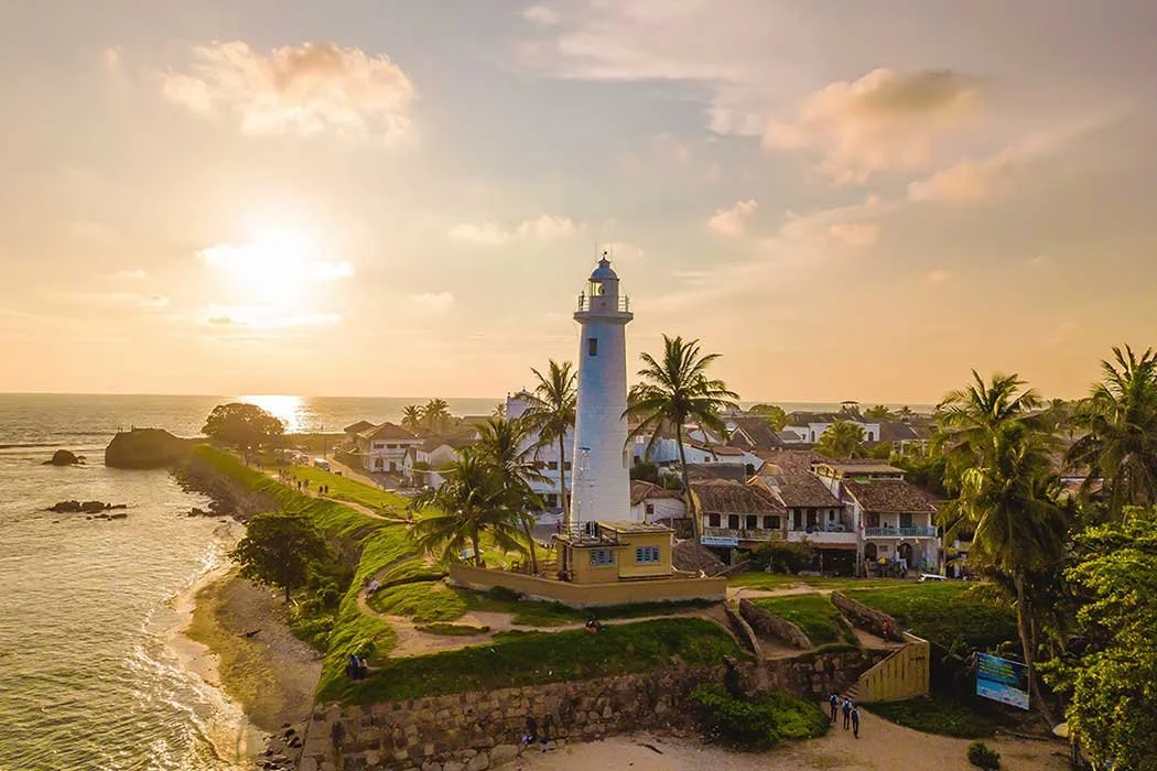 Lighthouse on a coastal hill with houses, palm trees, and a sunset over the ocean.