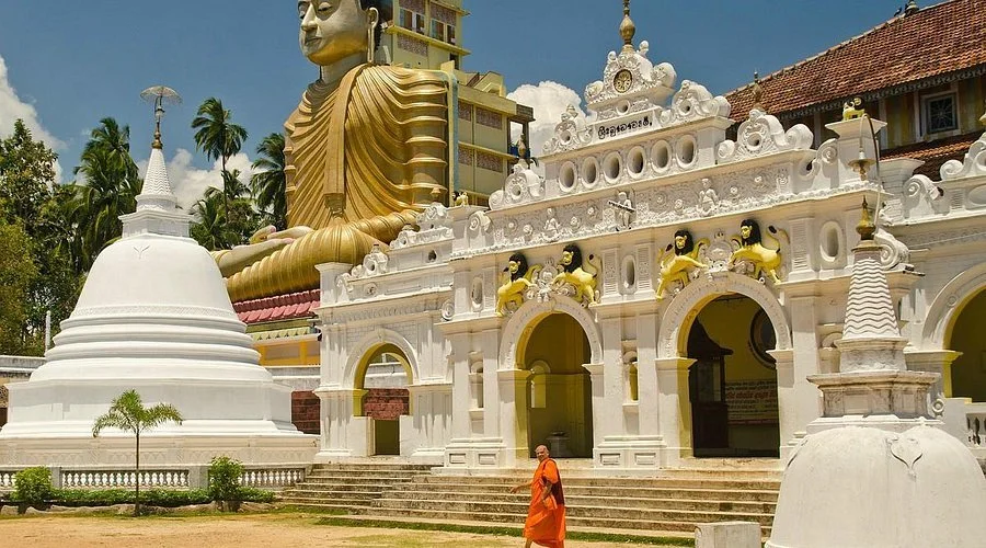 A large golden Buddha statue sitting outdoors behind a white temple with ornate decoration and smaller figures, with a monk walking nearby.