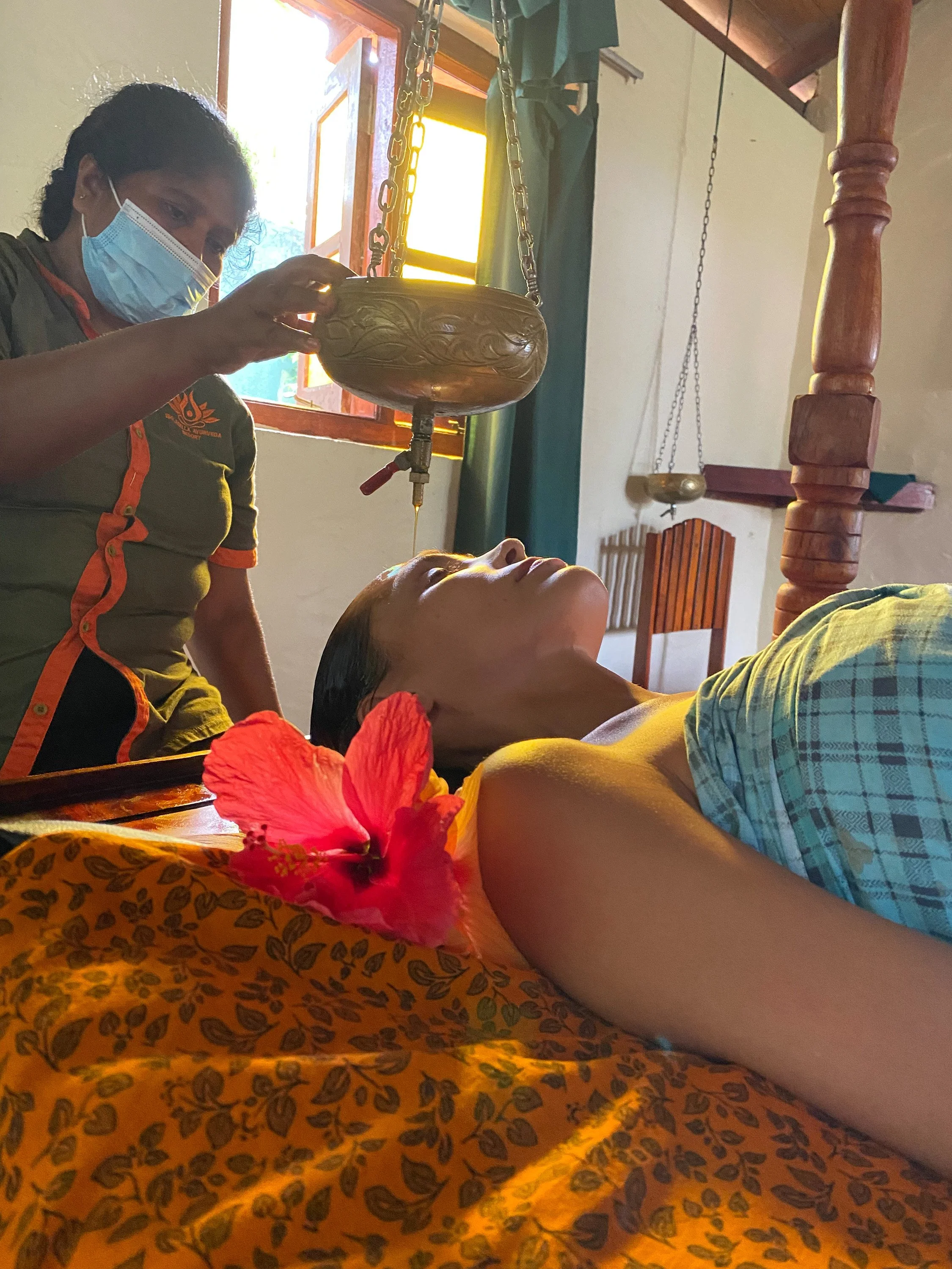 Žena leží pod oranžovou květinovou dekou a podstupuje bylinnou kosmetickou péči od odbornice, v místnosti se slunečním světlem – inspirace pro ájurvédský wellness pobyt a relaxační péči o pleť.