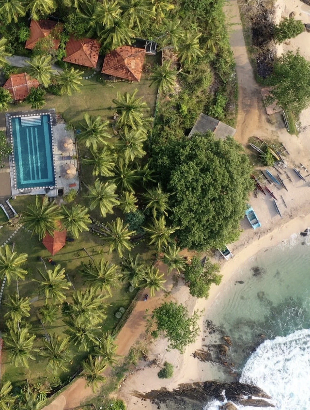 Letecký pohled na tropický resort u pláže s bazénem, palmami, malými budovami s červenými střechami, loděmi na pláži a mořskými vlnami – ideální inspirace pro ájurvédský wellness pobyt.