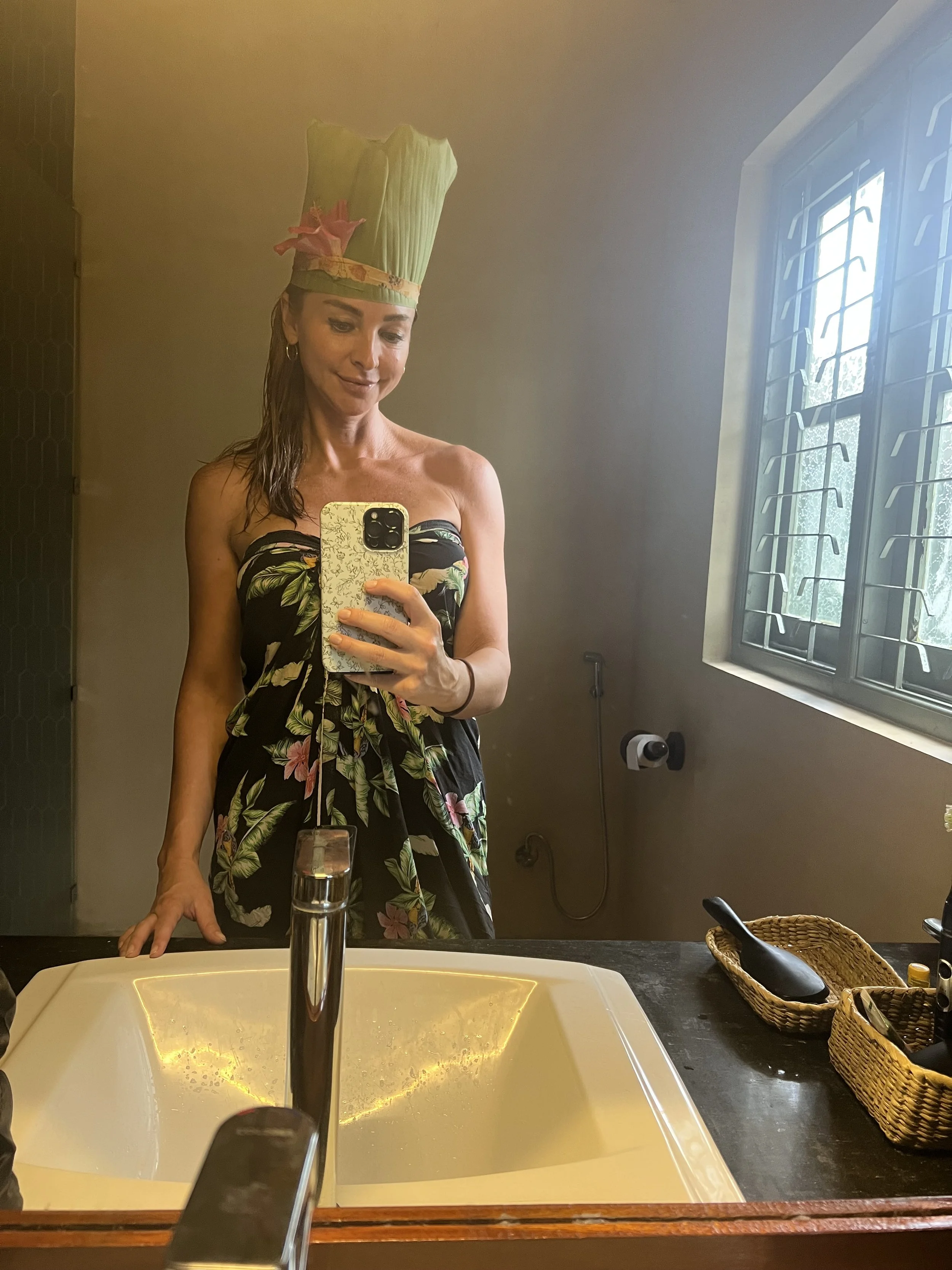 Žena si fotí selfie v koupelnovém zrcadle, má strapless šaty s květovaným vzorem a vysoký dezertní klobouk s růžovými dekoracemi – inspirace pro exotické zážitky a ájurvédský wellness pobyt.