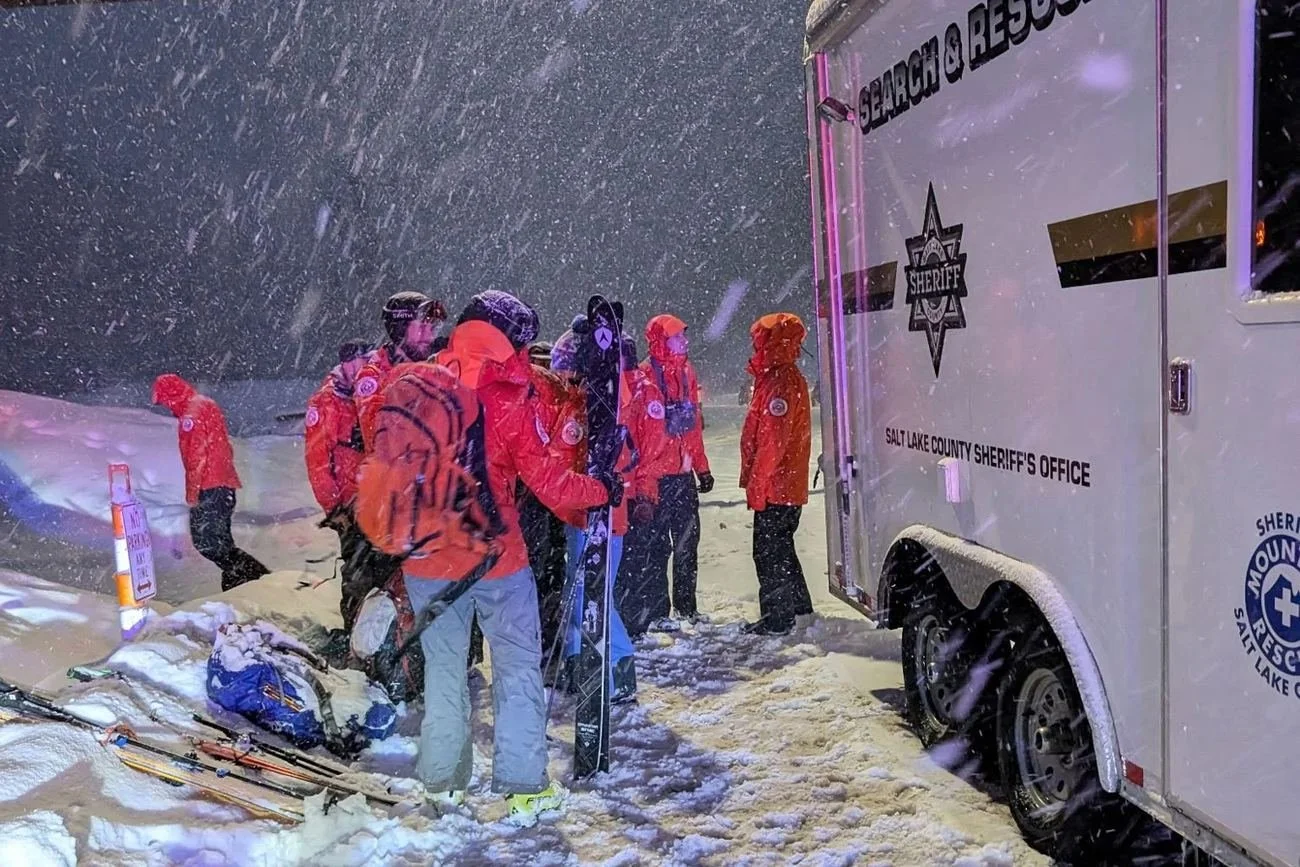 Search and rescue volunteers with skis beside the team trailer during heavy snowfall.