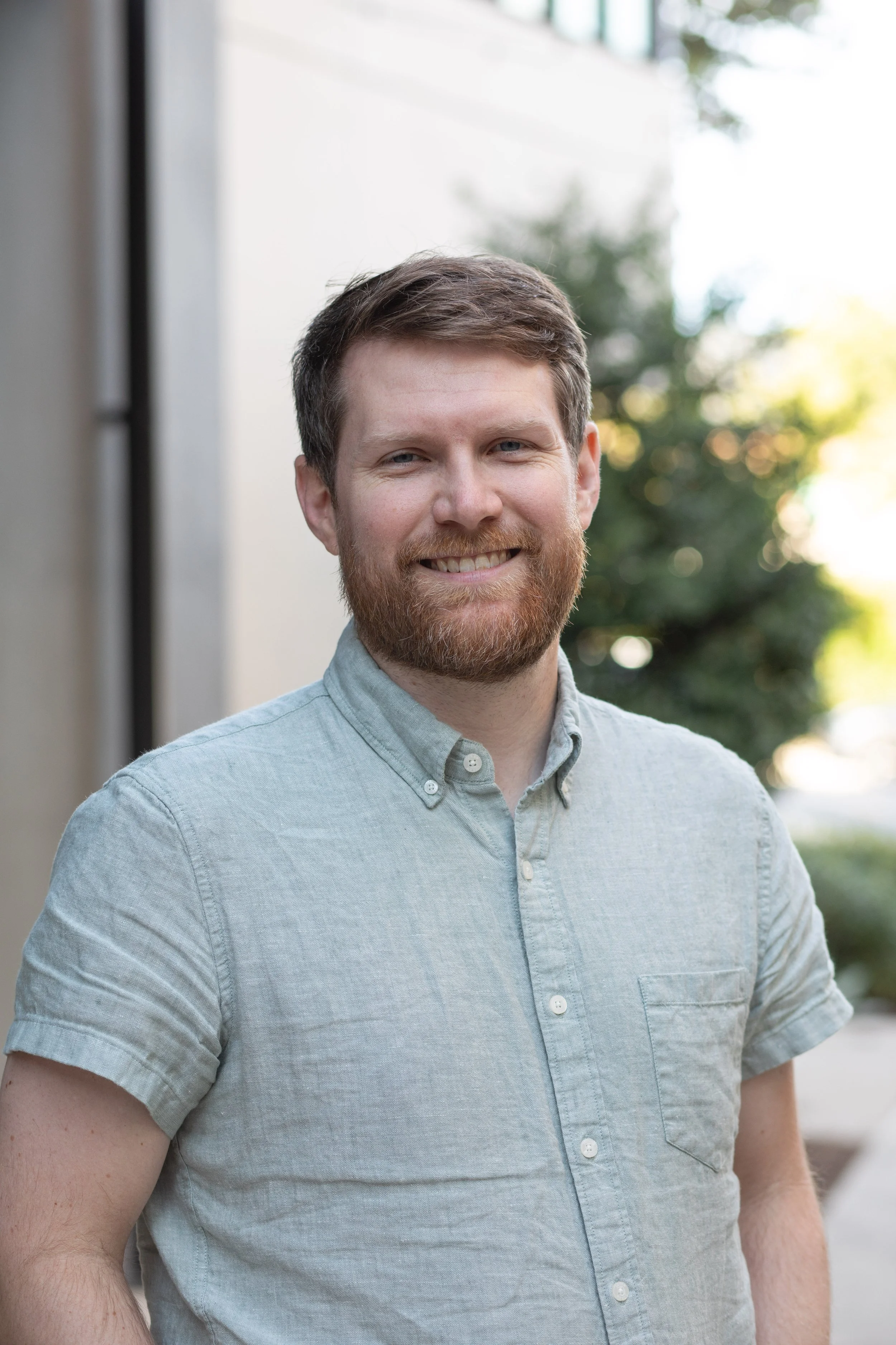 A smiling man with a beard and short hair, wearing a light-colored button-up shirt, standing outdoors with blurred trees and sunlight in the background.