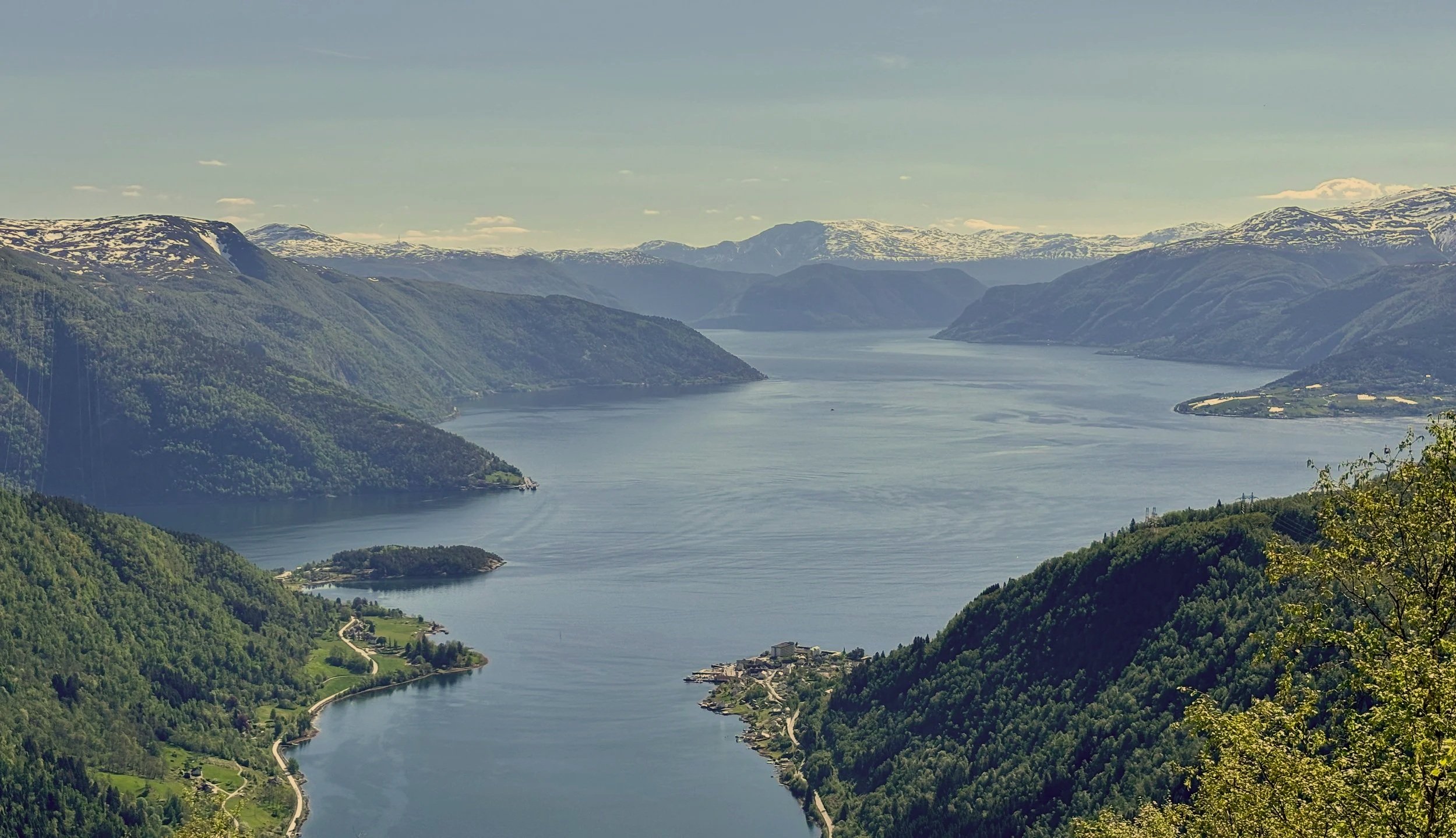 Scenic view of a fjord with steep, green mountains on both sides, snow-capped peaks in the background, and a small village near the water's edge.