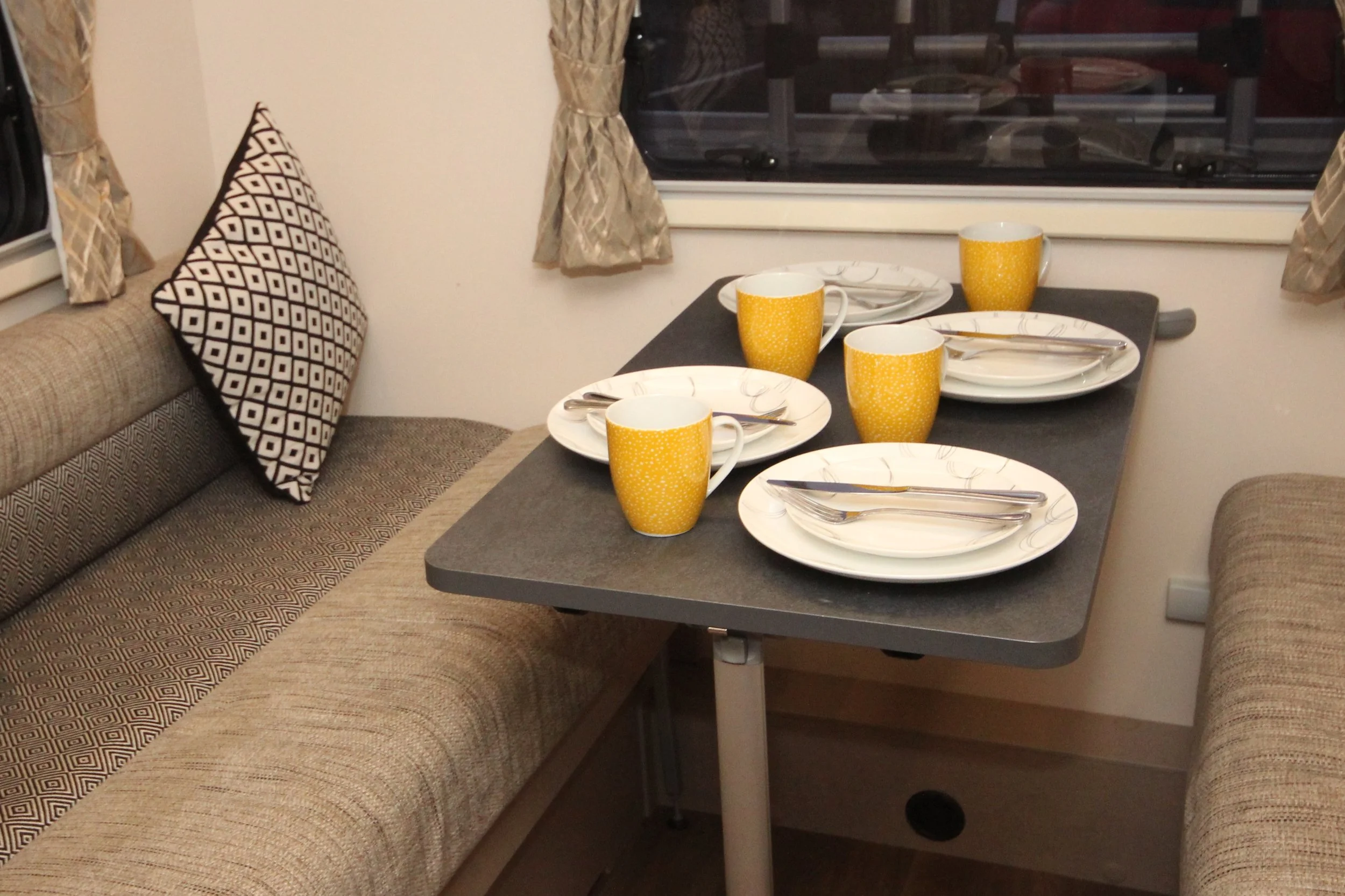 A table set for four with yellow cups, white plates, and silverware, surrounded by a beige cushioned bench and a beige wall with curtains, near a window.