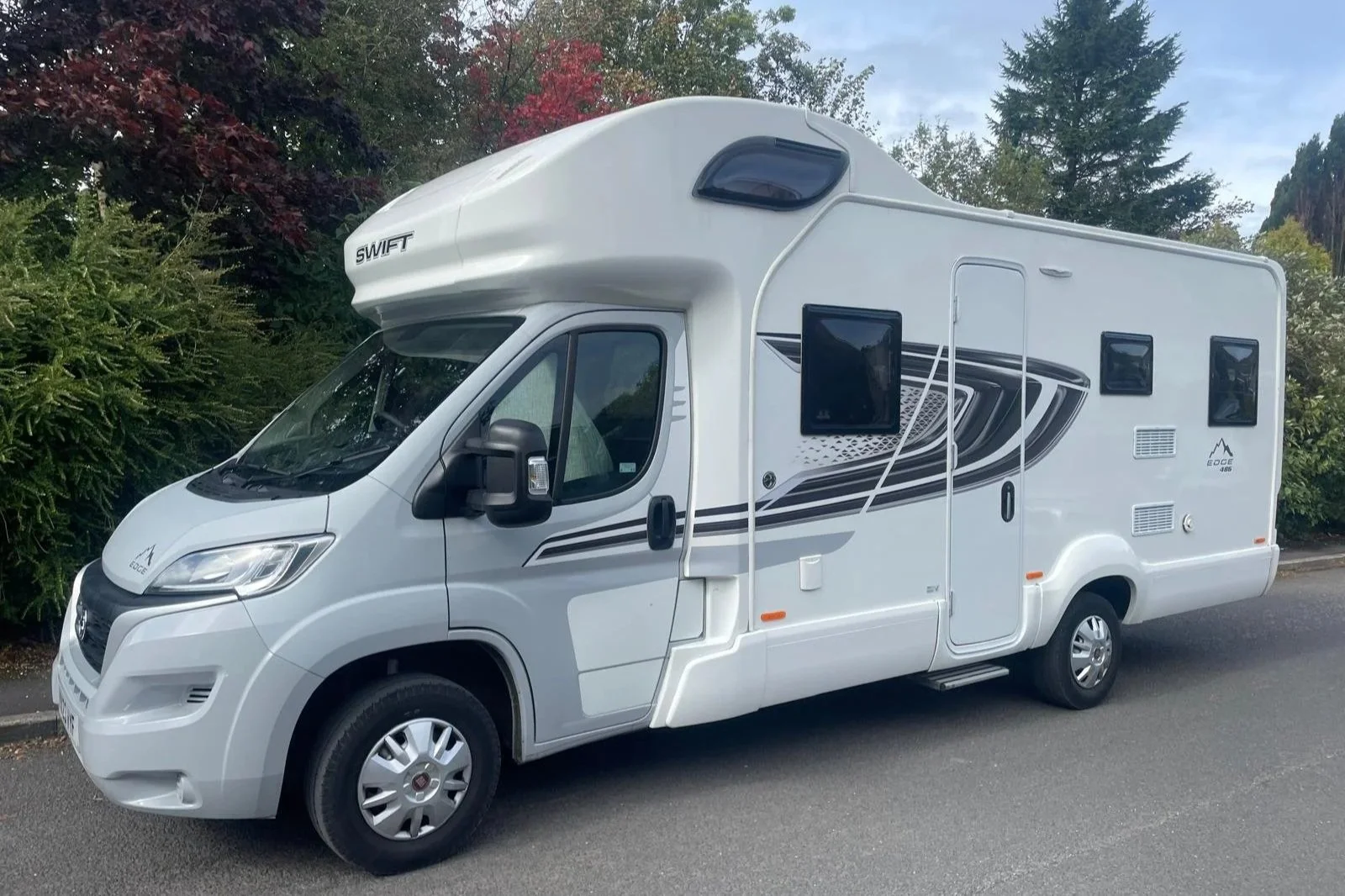 A white motorhome camper van parked on a street with trees and bushes in the background.