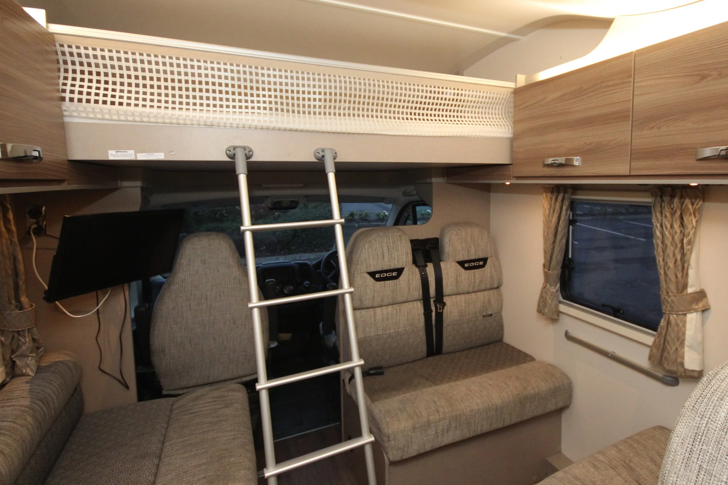 Interior view of a campervan showing seats, windows with curtains, an overhead bed with a ladder, and a TV mounted on the wall.