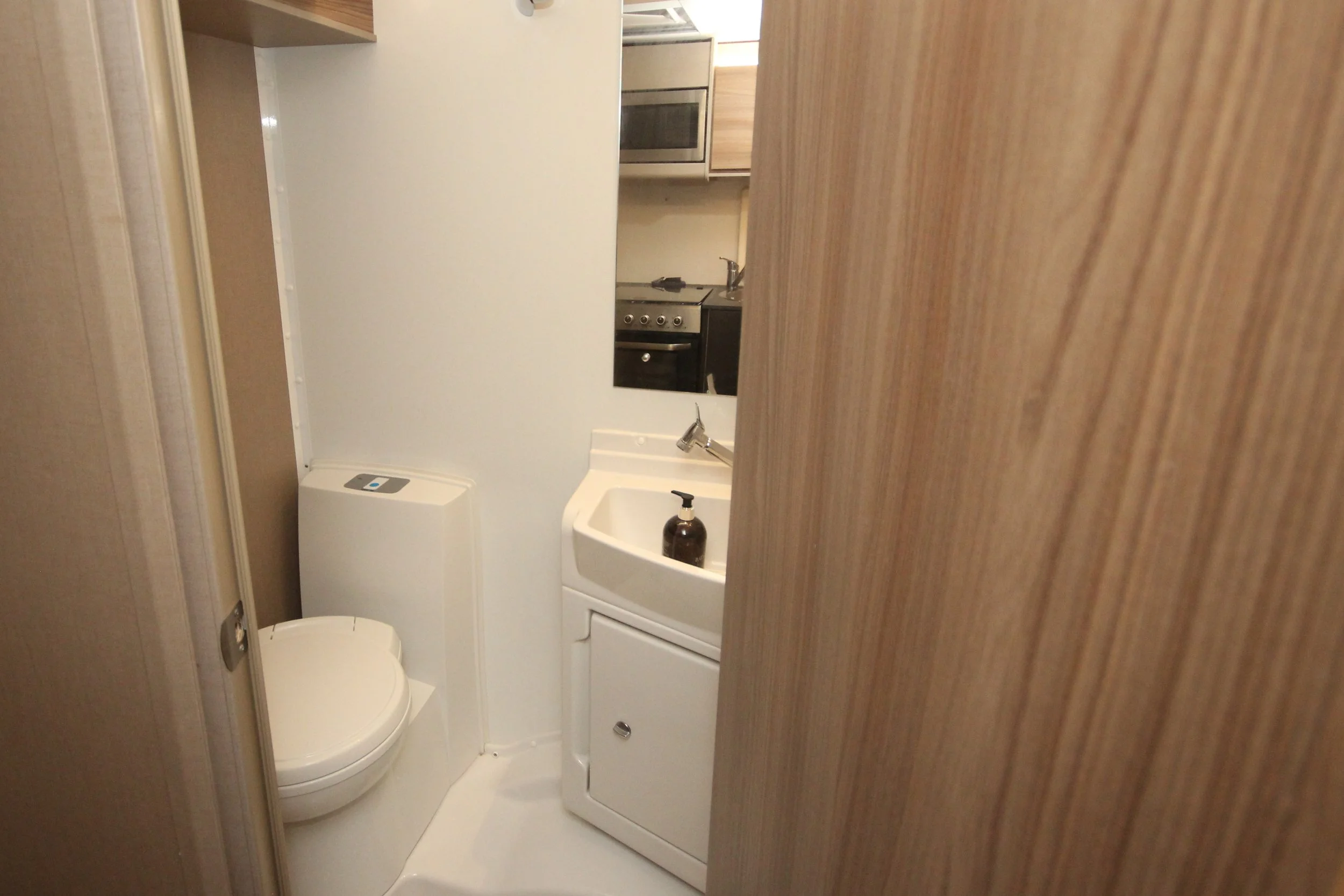 Small bathroom with a white toilet, a white sink with a brown soap dispenser, and a mirror above the sink. Kitchen appliances are visible through the doorway, including a stove and microwave. The room has beige and wooden accents.