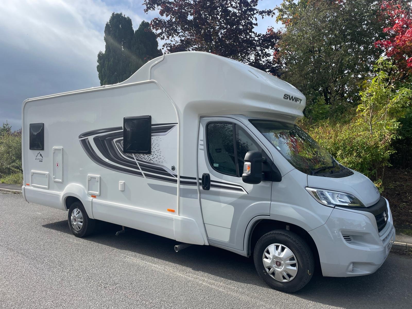 White 2023 motorhome with black and gray graphic decals, pitched on a parking lot with trees and colorful foliage in the background.