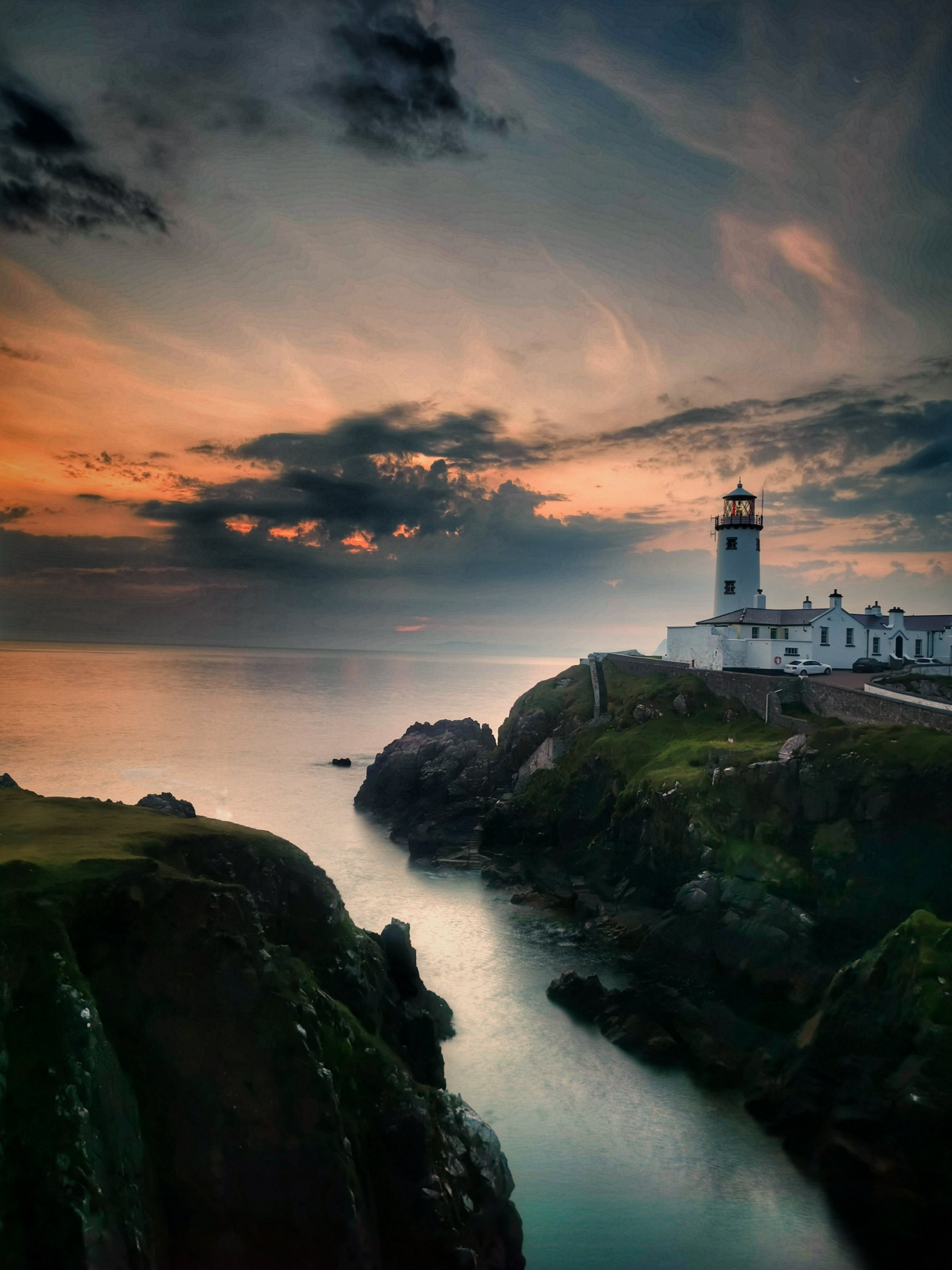 Fanad Lighthouse, Donegal, Ireland. A lighthouse on a rocky cliff overlooking the ocean at sunset with cloudy sky.