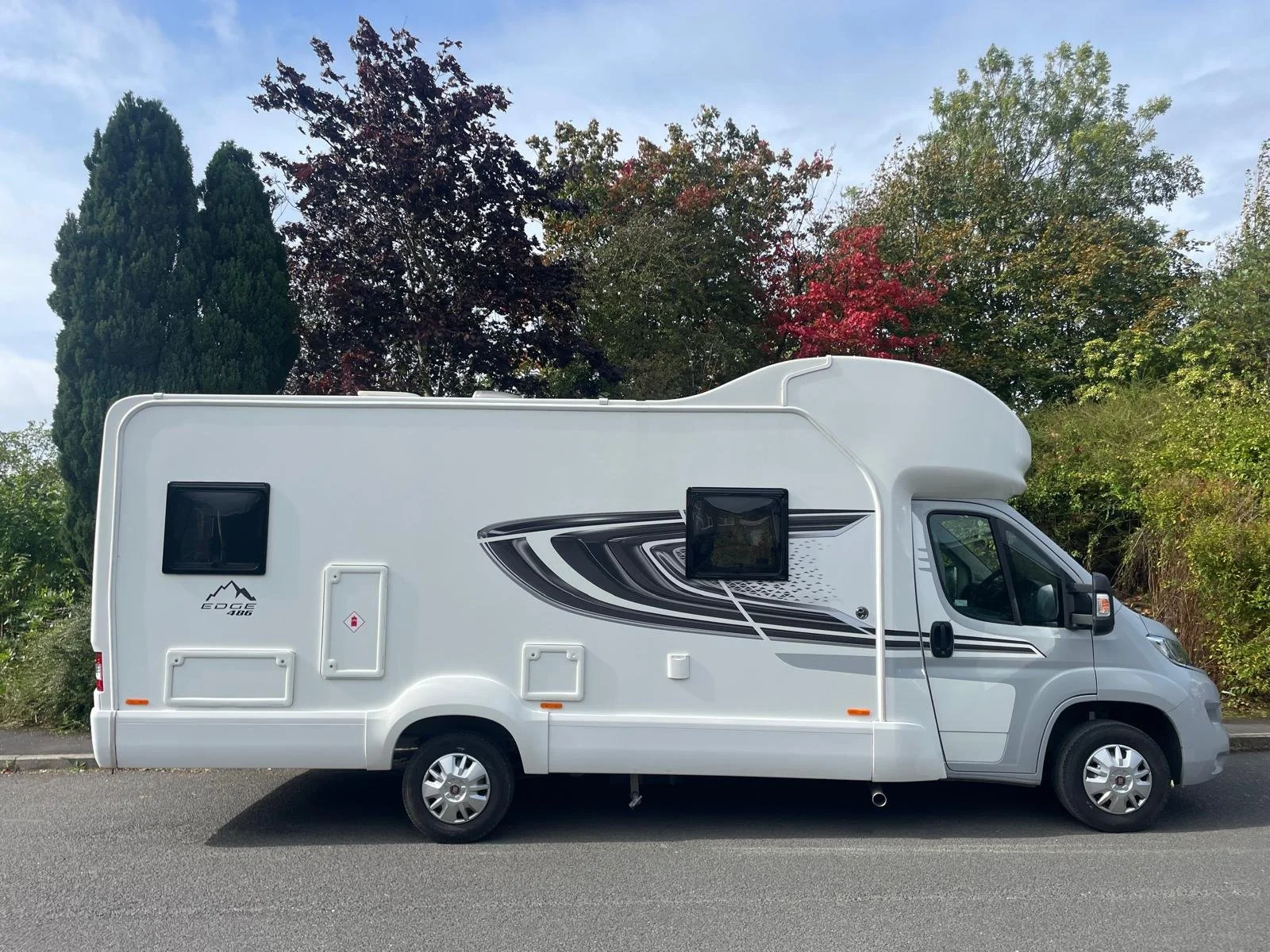 White motorhome parked on the street with trees with fall foliage in the background.