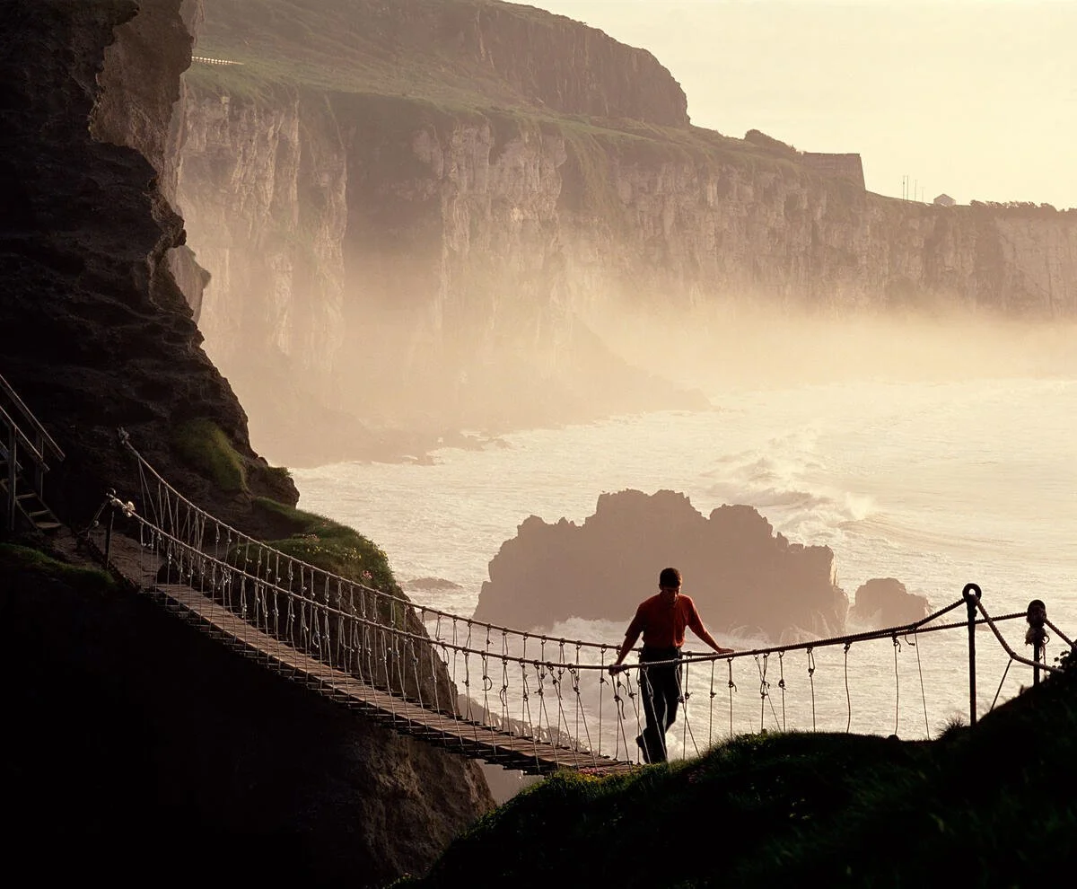 Carrick-A-Rede Rope Bridge, North Coast, Northern Ireland. A person walking on a suspension bridge along a rocky coastline with cliffs and ocean waves, during sunset or sunrise.