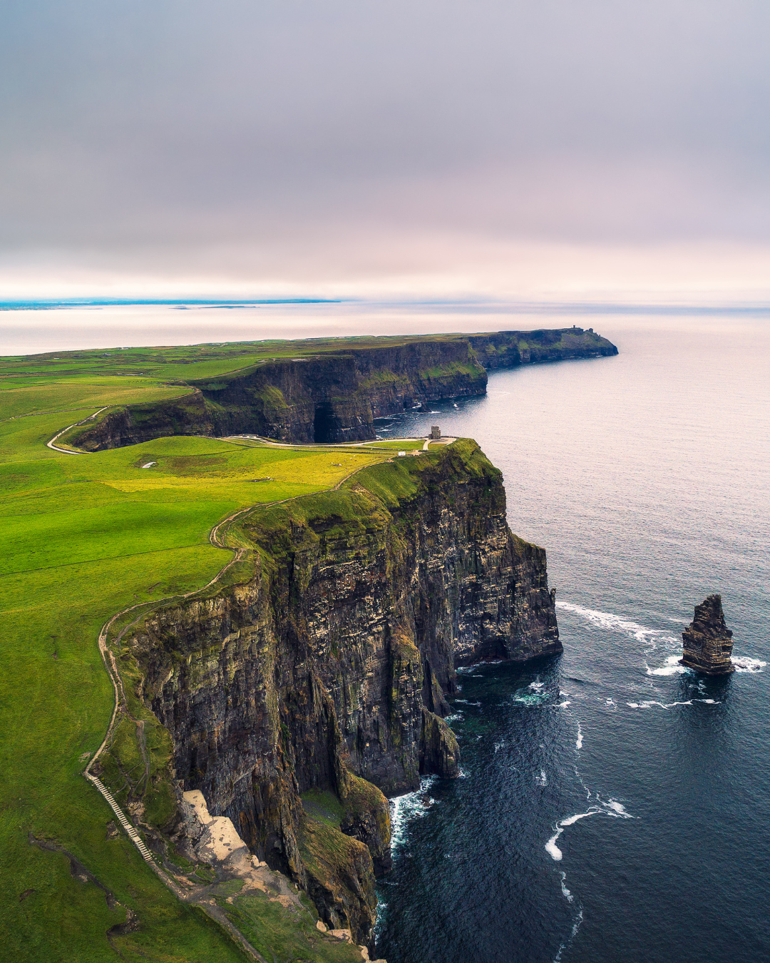 Cliffs of Moher coastline with green grassy top, steep cliffs, and outcroppings in the Atlantic Ocean under cloudy sky.