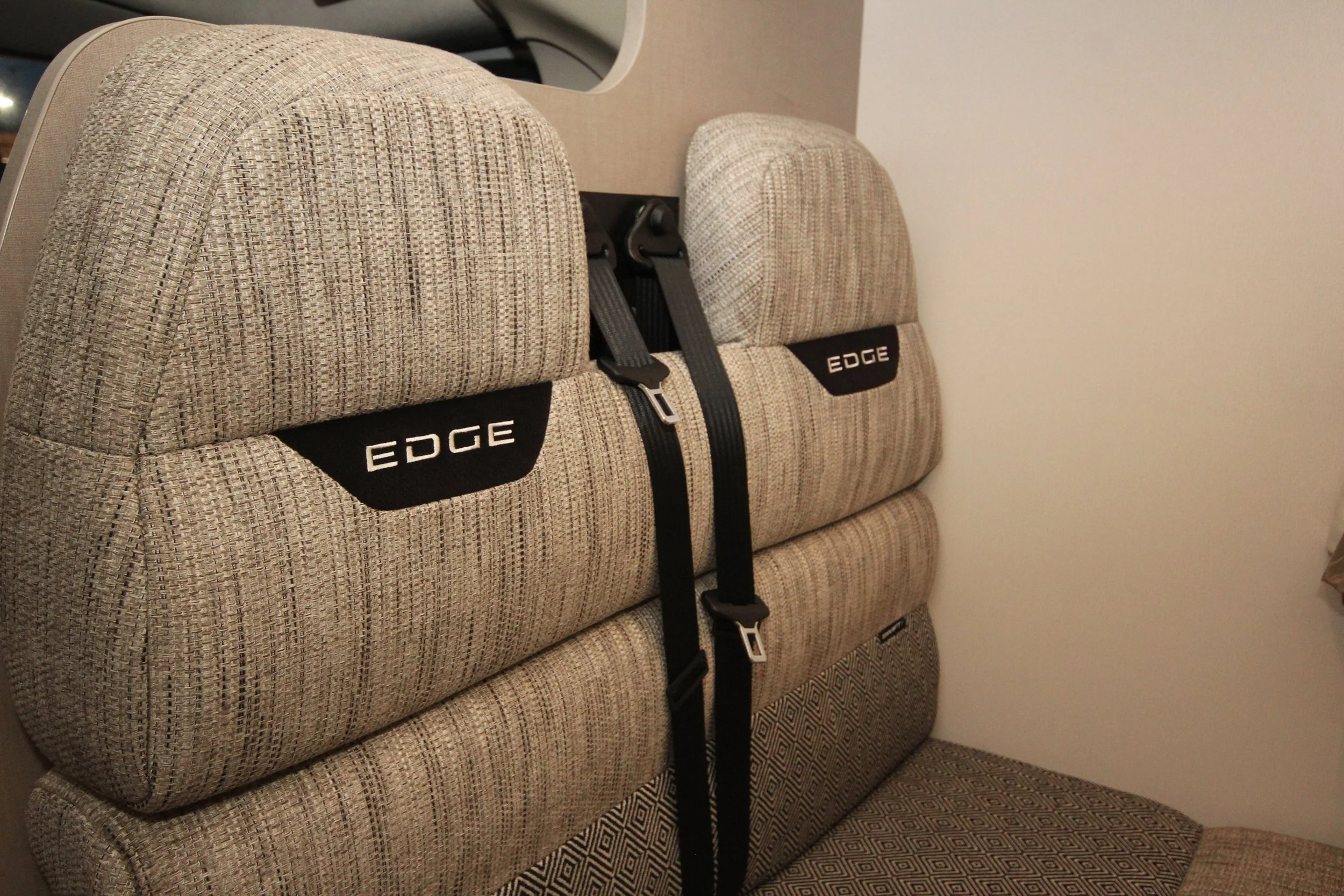 The image shows a beige headrest and backrest of a seat with a black strap hanging from it, labeled 'EDGE'. The seat and headrest are covered in a textured, patterned fabric, and the background shows a plain wall.
