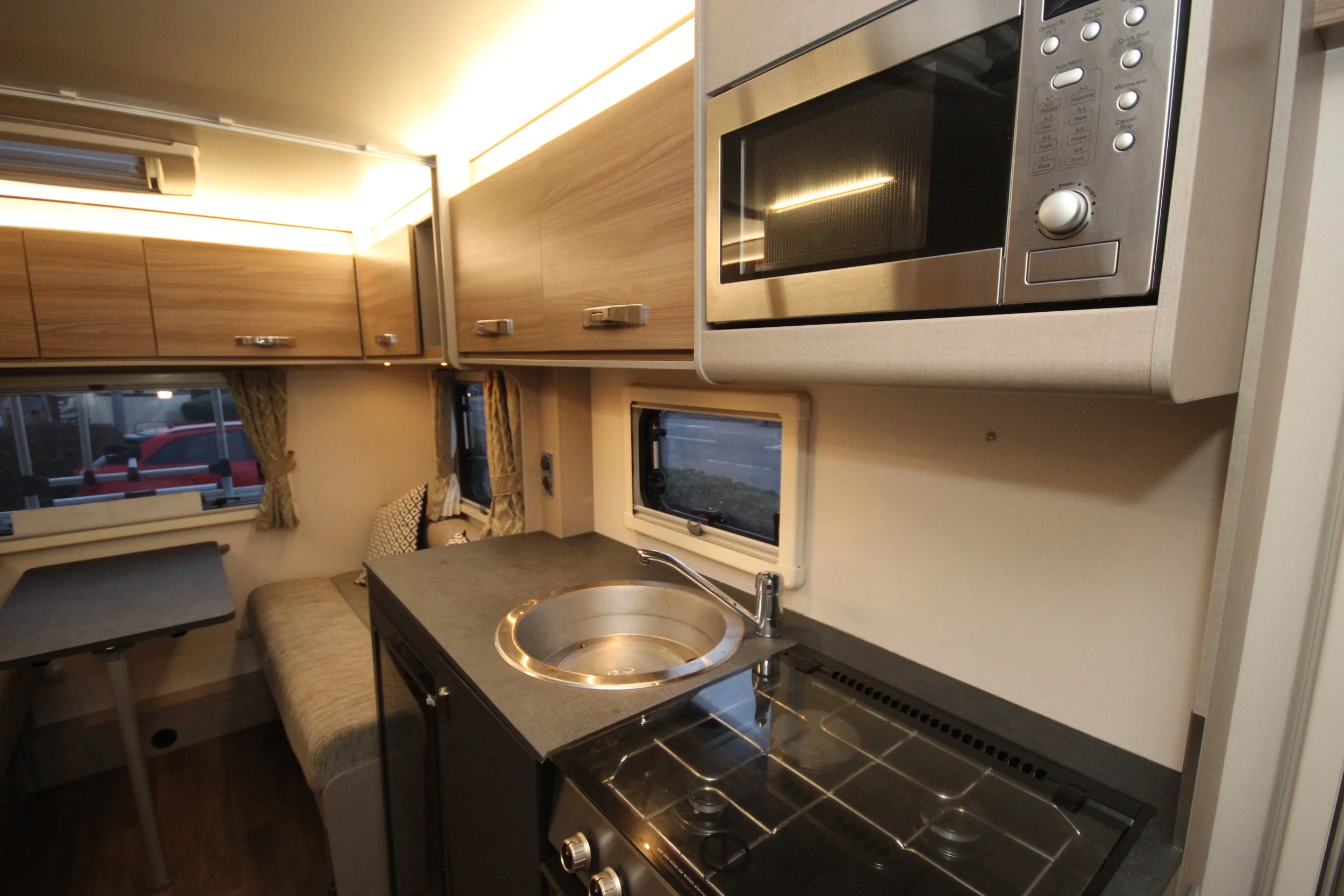 Interior of a compact camper van kitchen with a microwave, small sink, and stove, with a seating area and windows in the background.