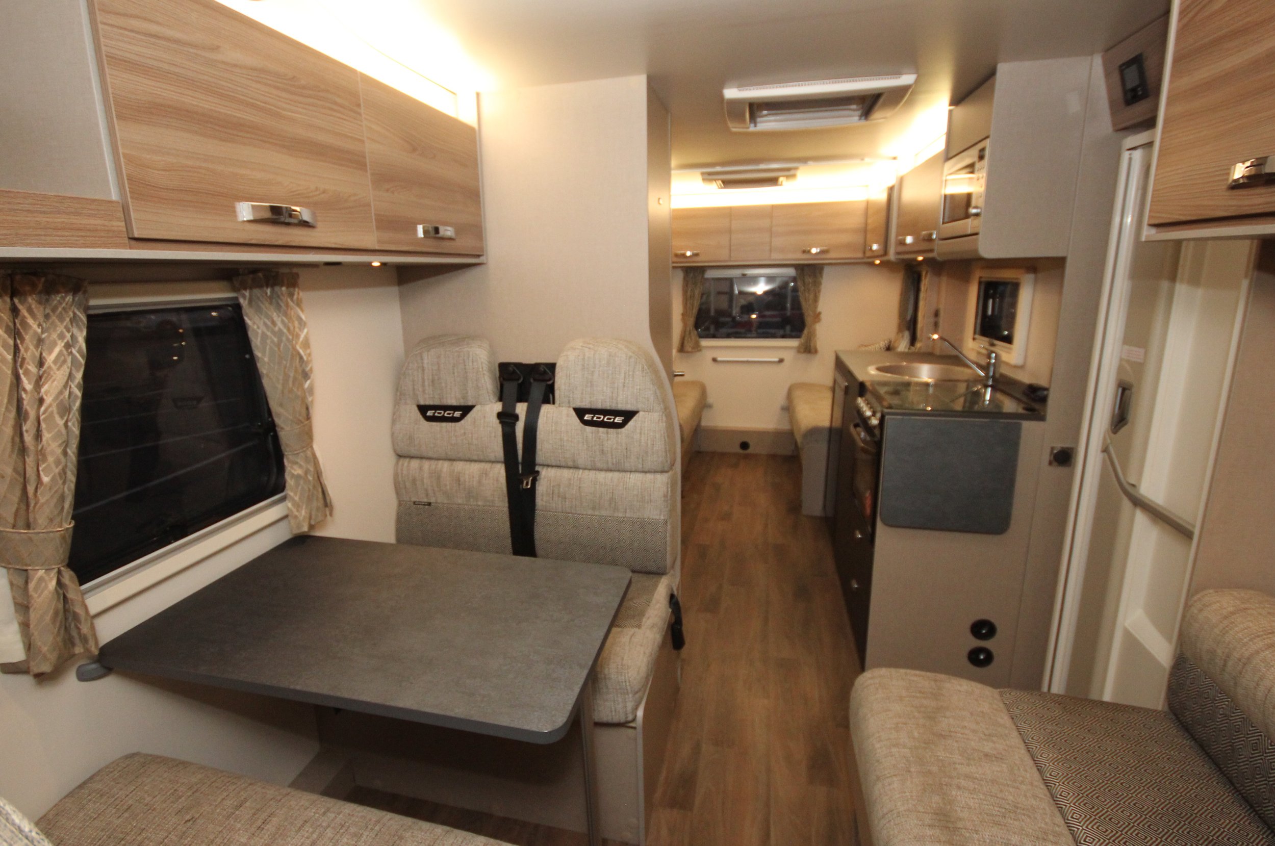 Interior of a compact camper with seating, a small dining table, a kitchen area with a sink and stove, cabinets, windows with curtains, and wood flooring.