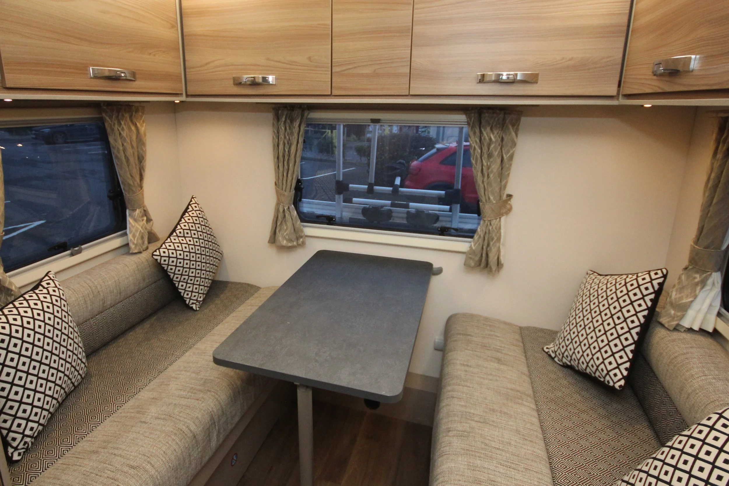 Inside a small RV with two cushioned bench seats, each with patterned pillows, surrounding a rectangular table near two windows with patterned curtains.