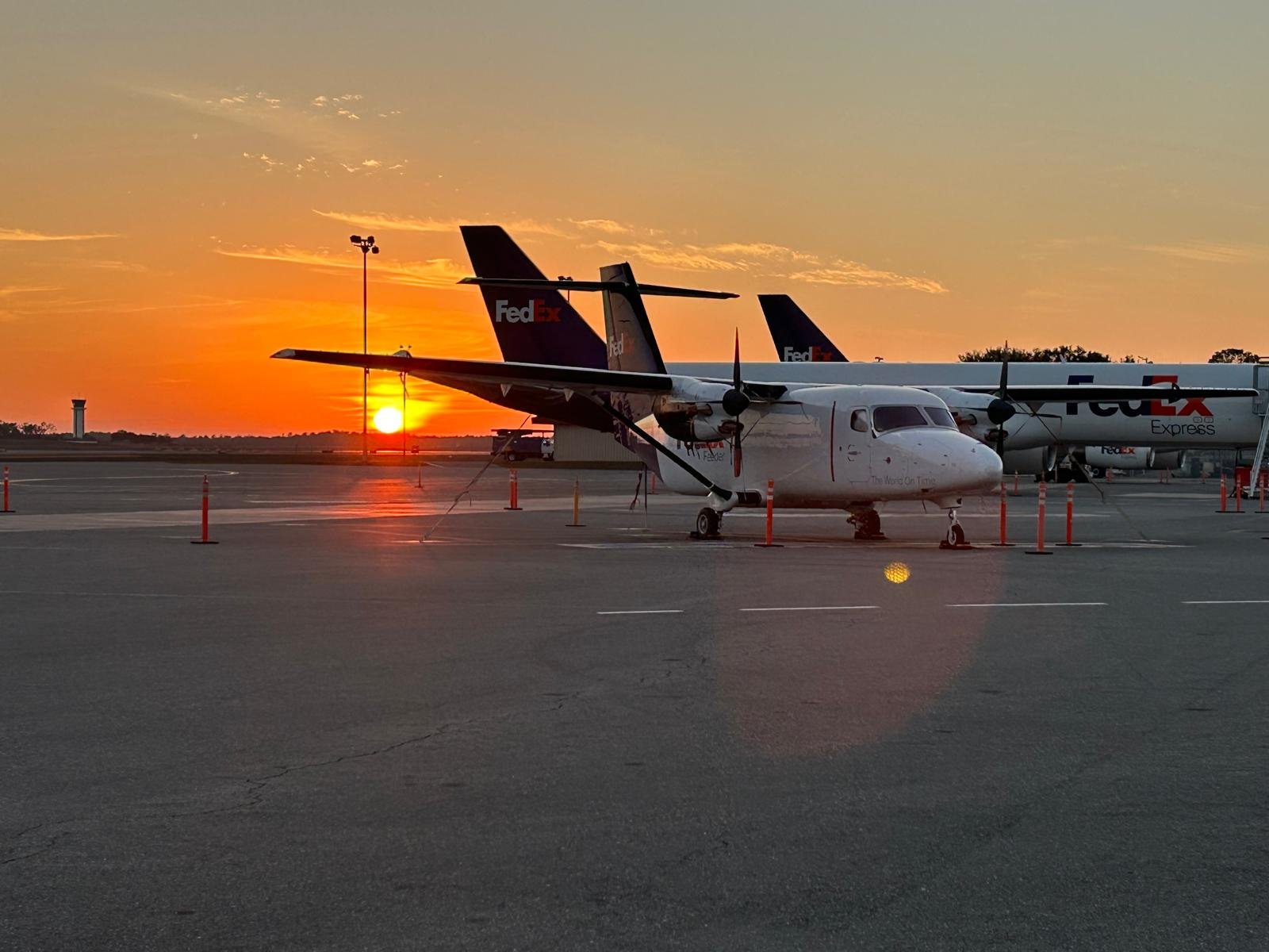 Propeller airplane with FedEx logos parked on tarmac at sunset with more FedEx planes in the background.