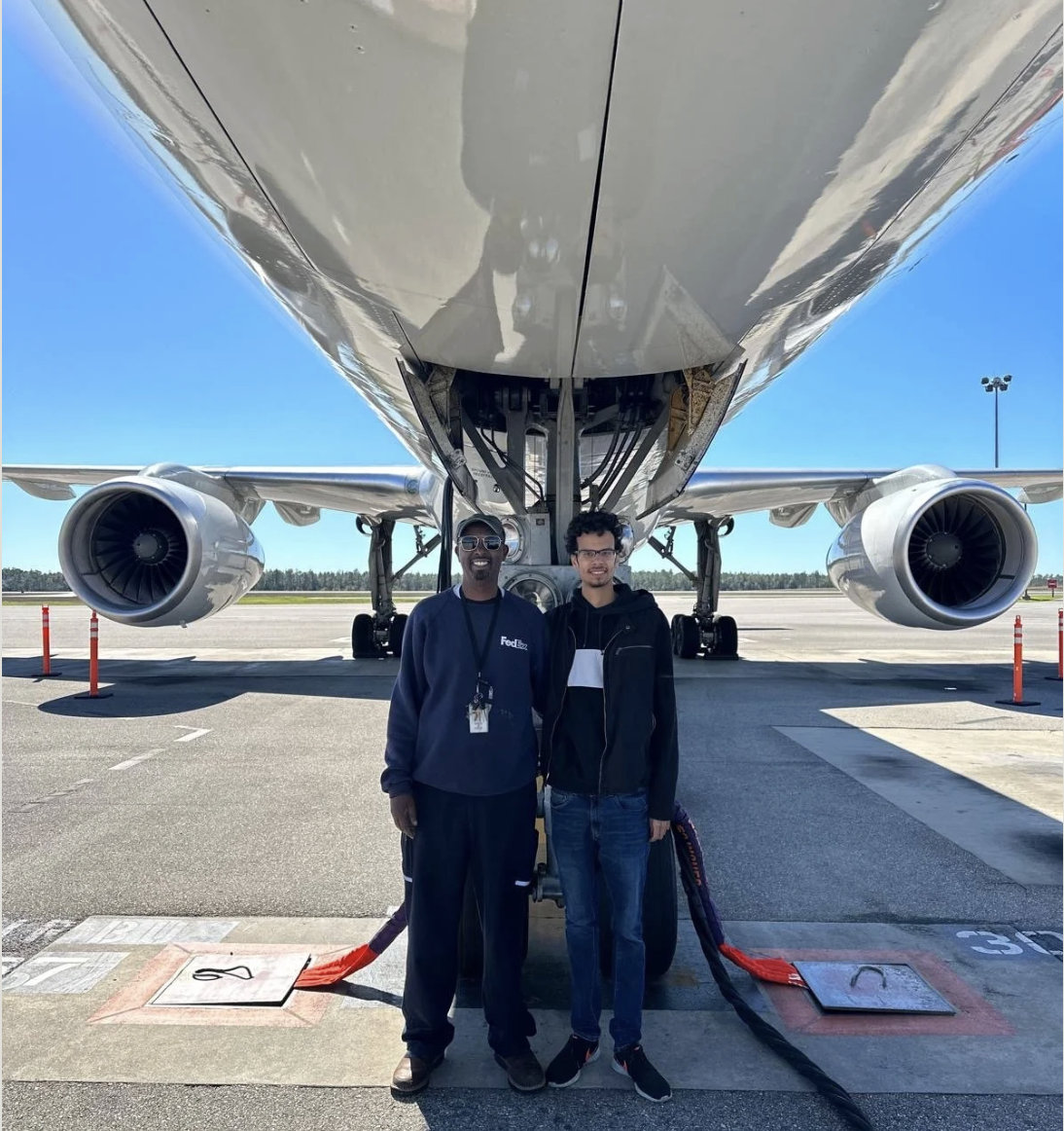 Two men standing in front of a large commercial airplane on the tarmac at an airport, with clear blue sky in the background.