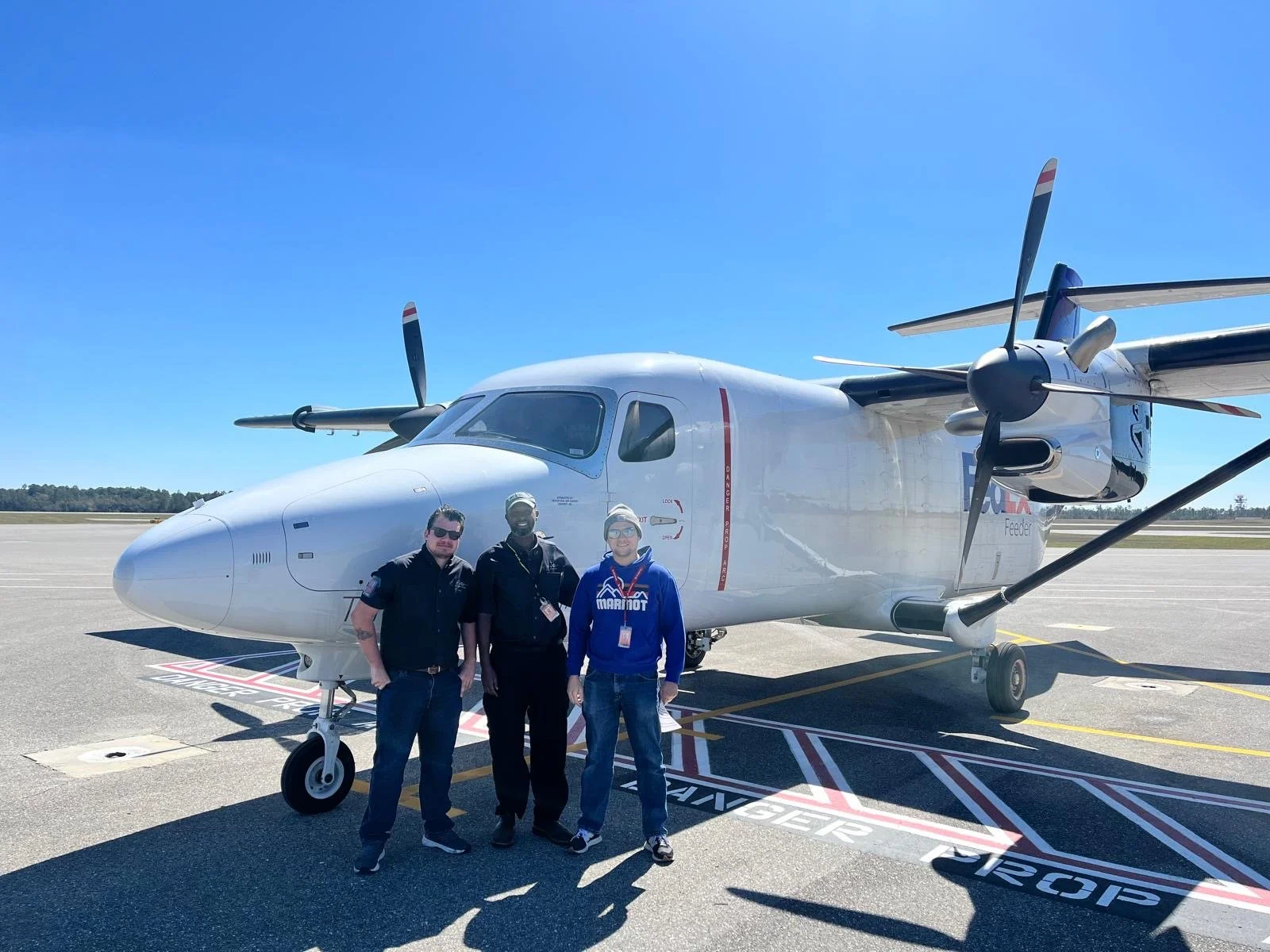 Three men standing in front of a white propeller aircraft on an airport tarmac under a clear blue sky.