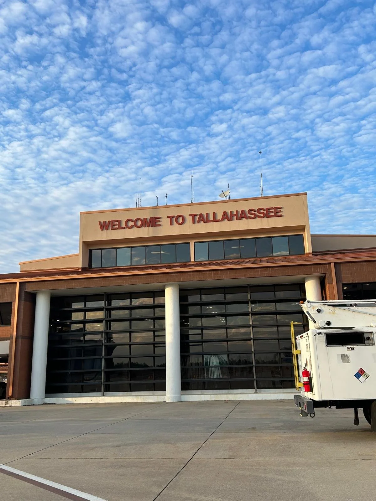The exterior of an airport terminal with a sign saying "Welcome to Tallahassee" in red letters, glass windows, and a cloudy sky above.
