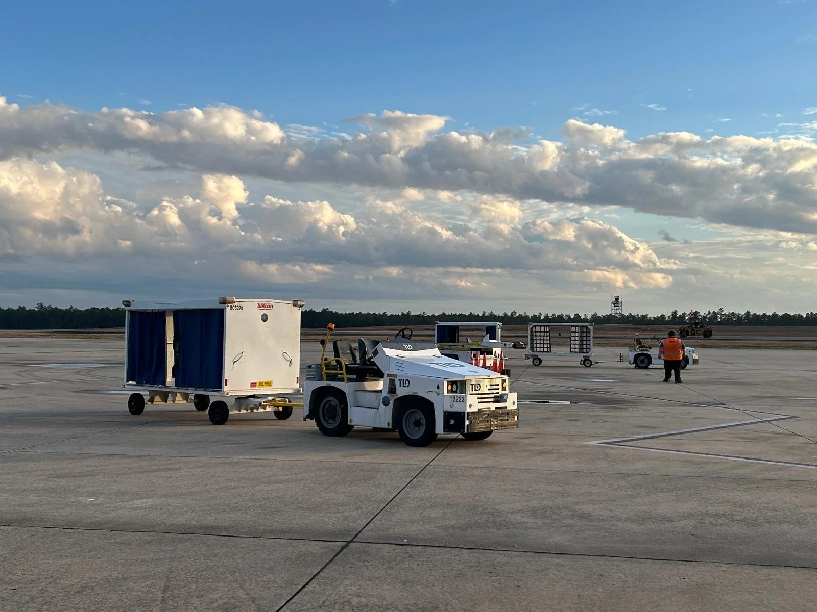 Airport tarmac with baggage carts, a tug vehicle, a ground crew member in orange vest, and a tractor in background under cloudy sky.