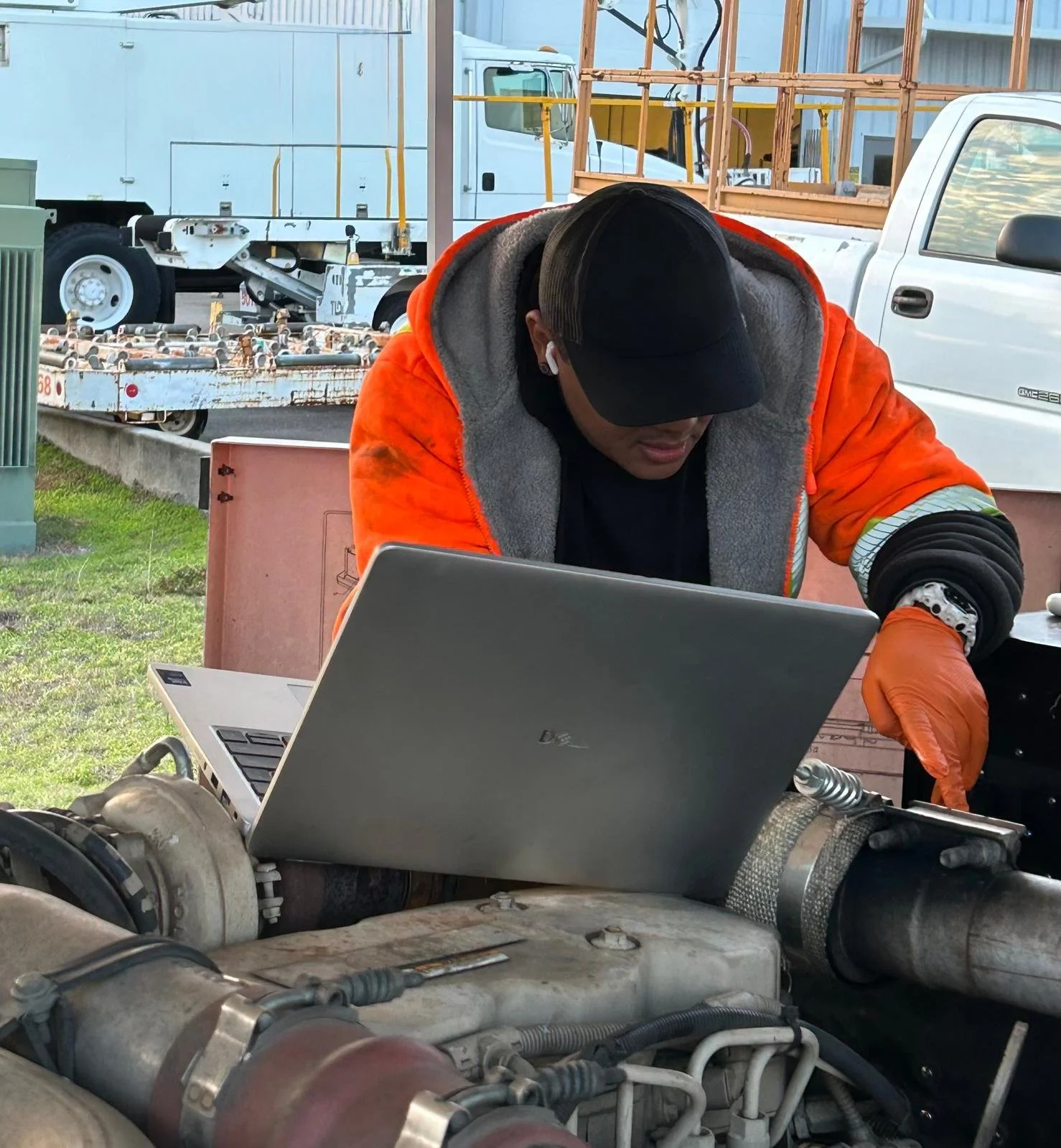 A man wearing an orange jacket and black cap working on an engine, using a laptop, outdoors with trucks and construction equipment in the background.
