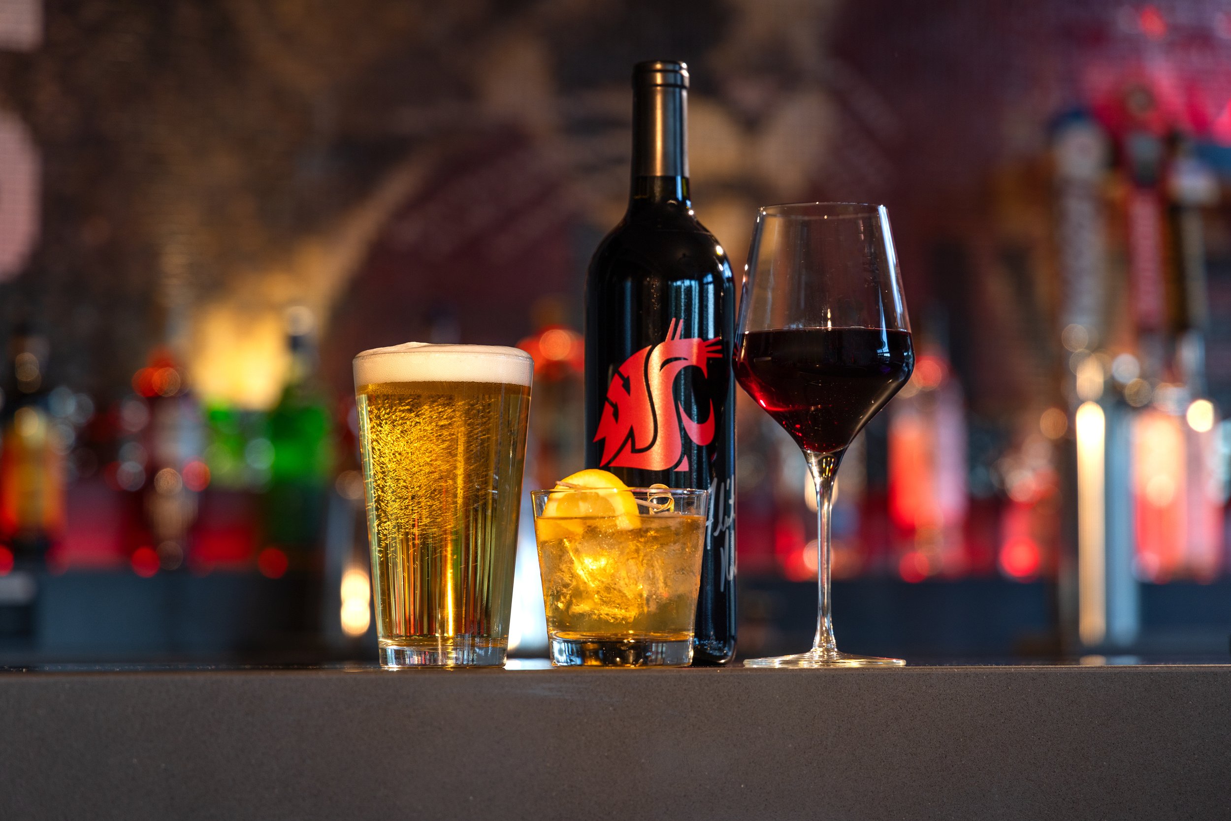 A pint glass of beer, a cocktail with lemon, a bottle of red wine, and a glass of red wine are on a bar counter with colorful blurred bottles in the background.