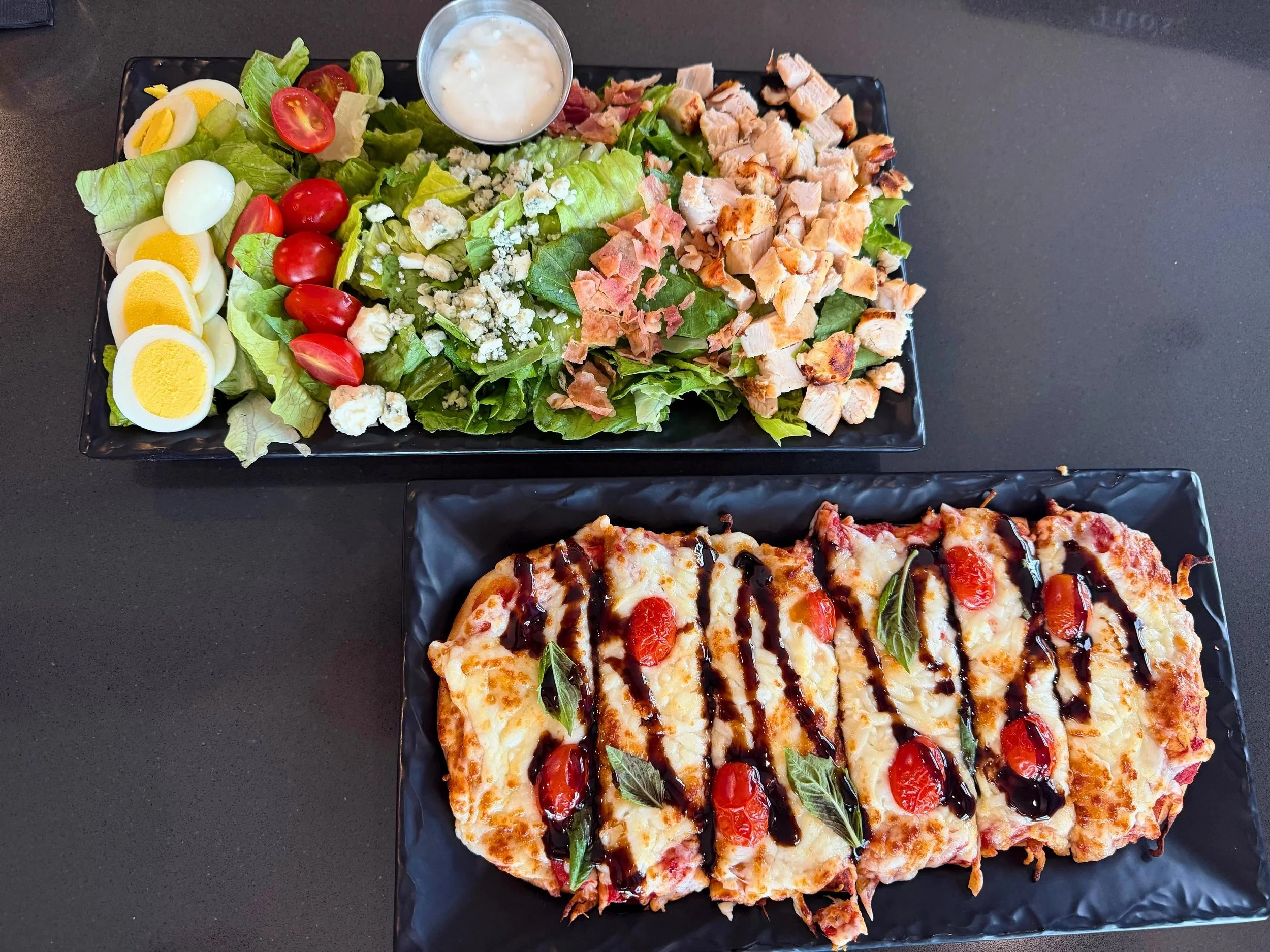 A black rectangular plate containing a slice of pizza with toppings including cherry tomatoes, basil, and mozzarella cheese, drizzled with balsamic glaze.