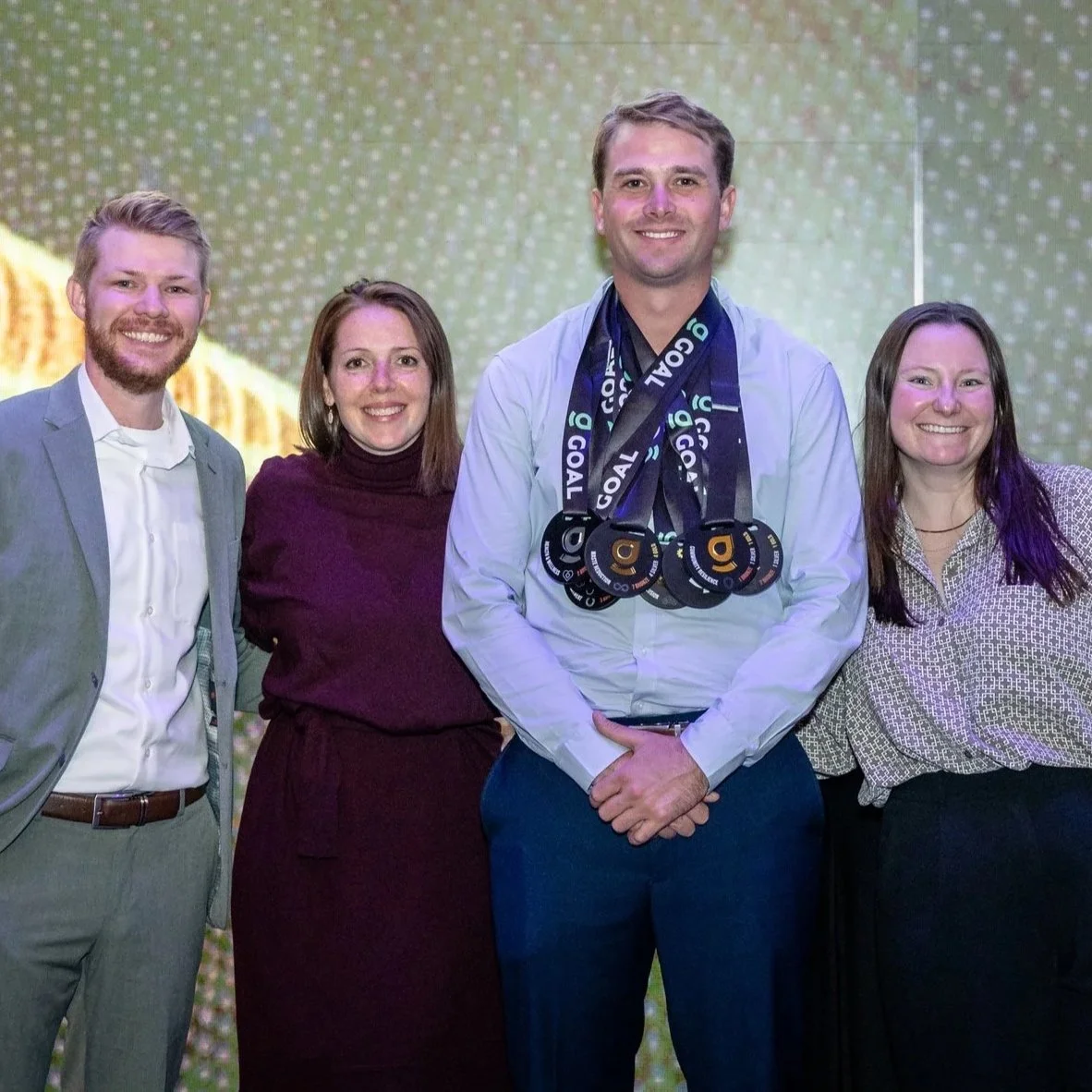 A group of four people standing together, celebrating at an event. The man in the center is wearing multiple medals around his neck.