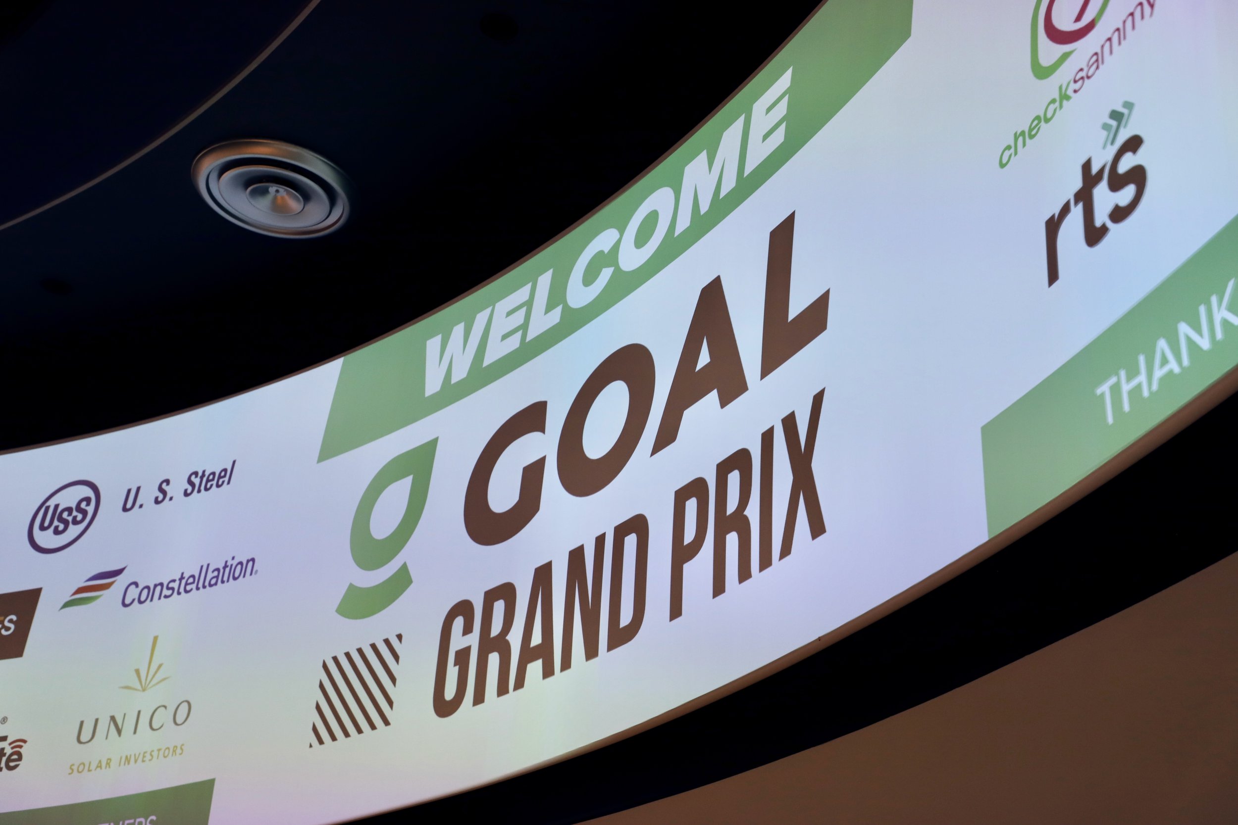 Large LED screen displaying a welcome message for the Goqal Grand Prix event, with sponsor logos including U. S. Steel, Constellation, UNICO Solar Investors, and checkSammy, in an indoor setting.