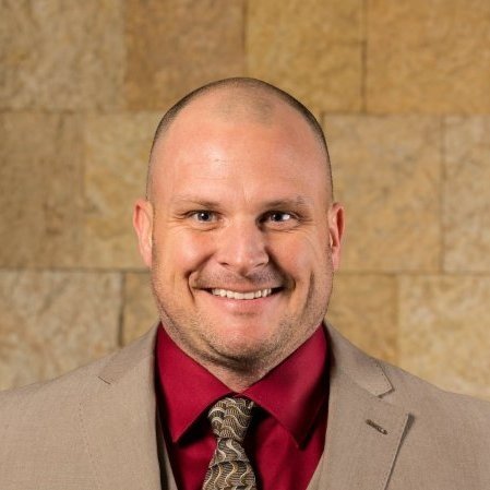 A man with a shaved head wearing a beige suit, red shirt, and patterned tie, smiling in front of a tan, brick wall background.