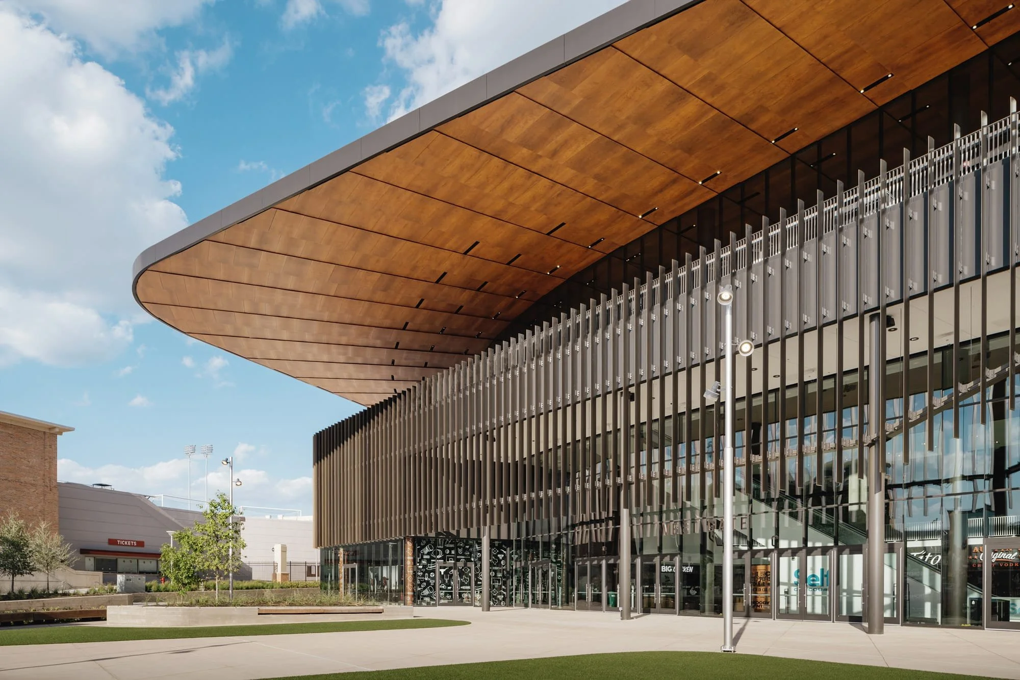 Modern building with a curved wooden overhang, glass Doors, and vertical metal slats, exterior of a sports arena or mall.