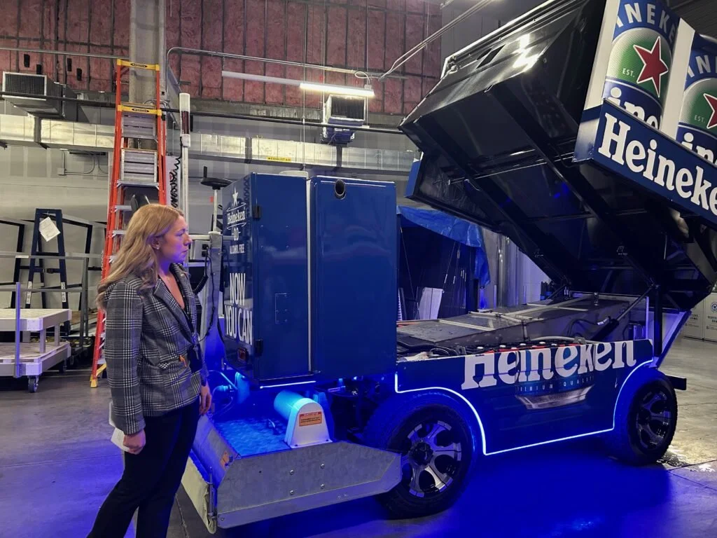 A woman with blond hair wearing a plaid blazer and black pants stands next to a blue promotional cart with Heineken branding in a warehouse-like setting.