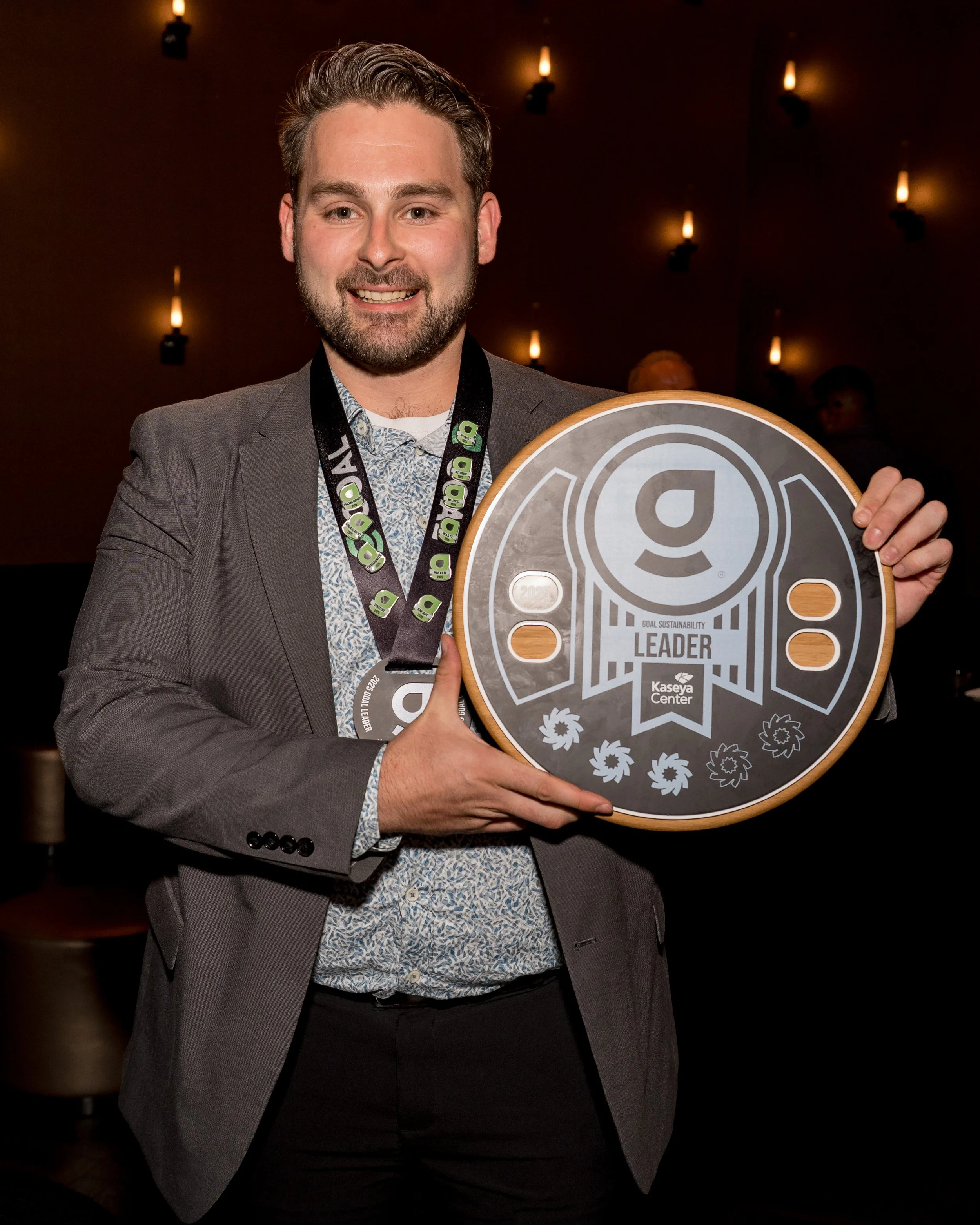 A man in a suit holding an award plaque that says 'Goal Sustainability Leader' at an event with dim lighting and hanging lamps.