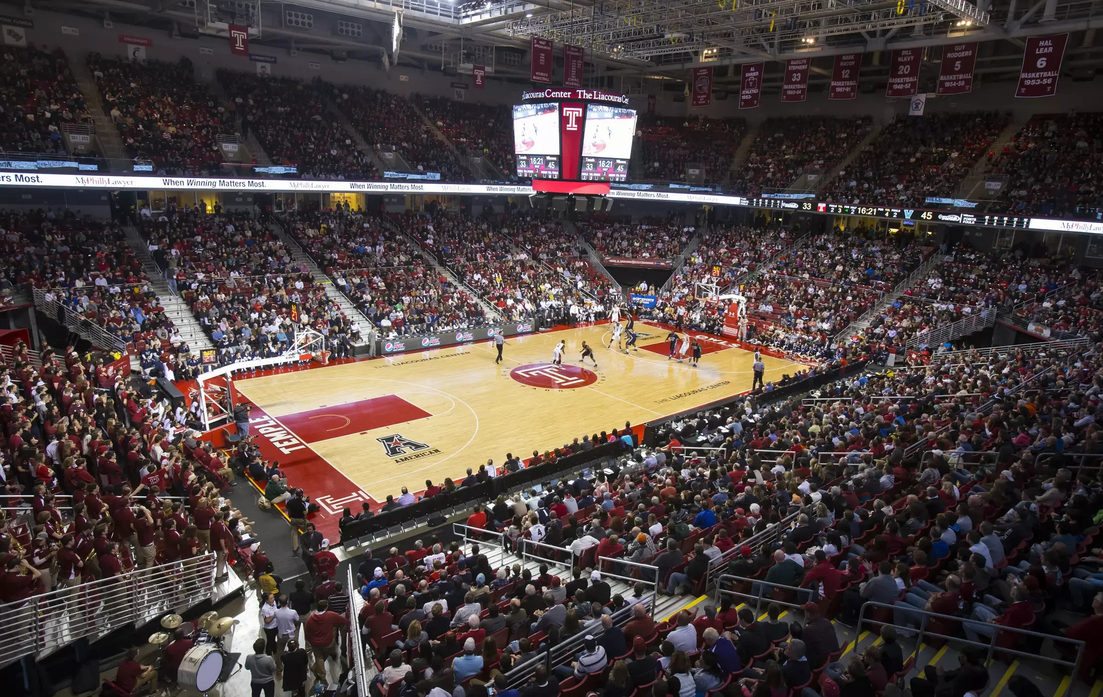 An indoor basketball game taking place at a large arena filled with spectators. The basketball court has players active, with a score display on an overhead screen showing 33 to 45 points.