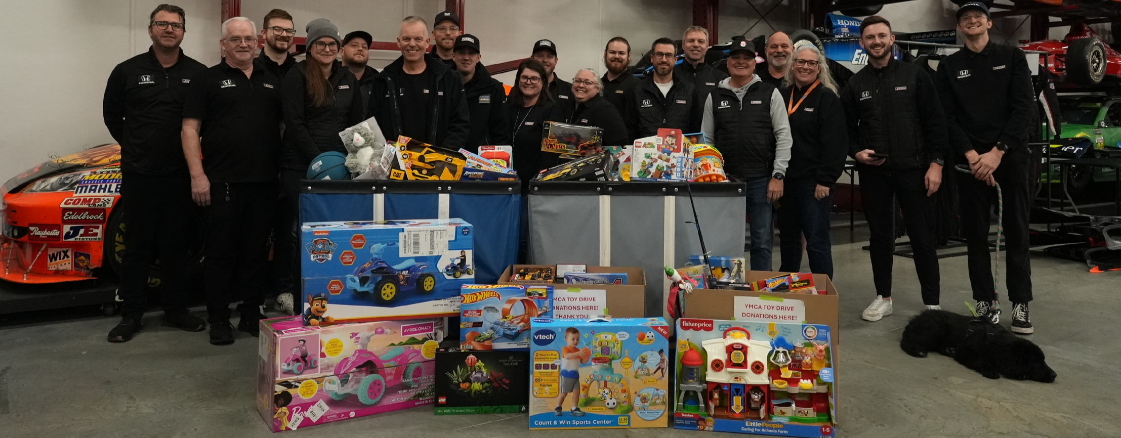 A group of people standing behind donation bins filled with toys for a toy drive event. They are indoors, with race cars visible on the sides, and a black dog lying on the floor nearby.