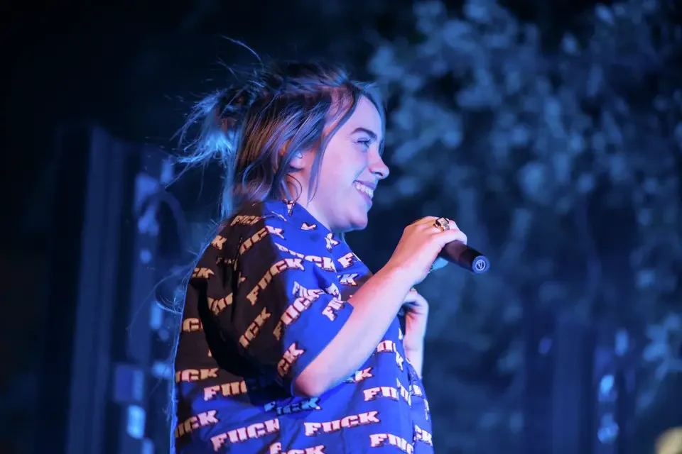 A young woman singing into a microphone on stage, smiling with her hair in a messy bun, wearing a blue T-shirt with goggles printed on it, with a blurred outdoor background.