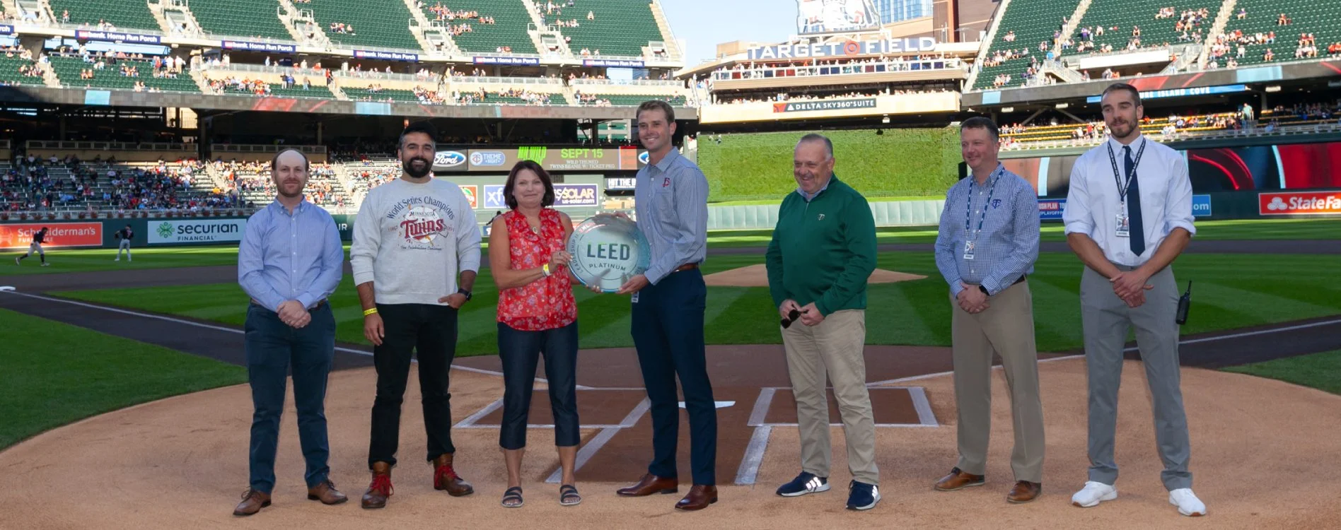 Group of seven people standing on a baseball field in a stadium, with large digital screen and seating in the background, during a stadium event.