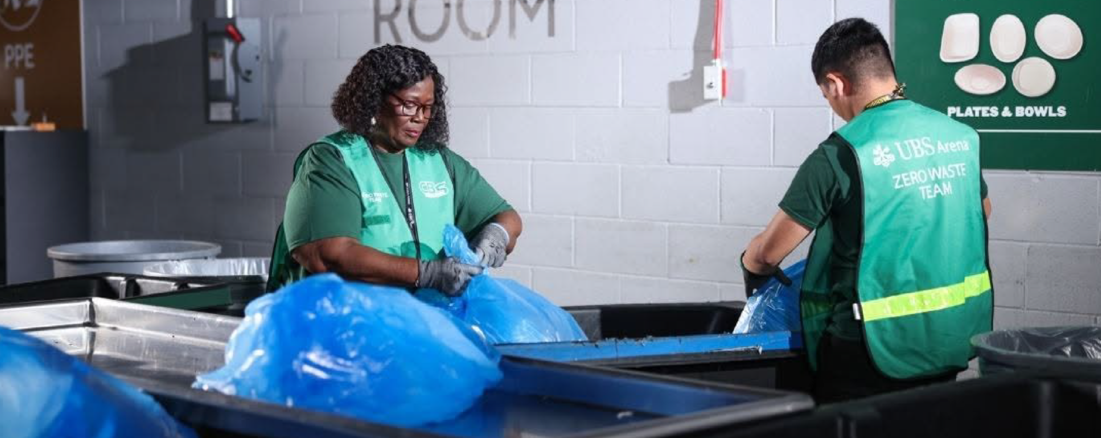 Two workers sorting recyclable materials in a waste management facility, wearing green shirts and gloves.