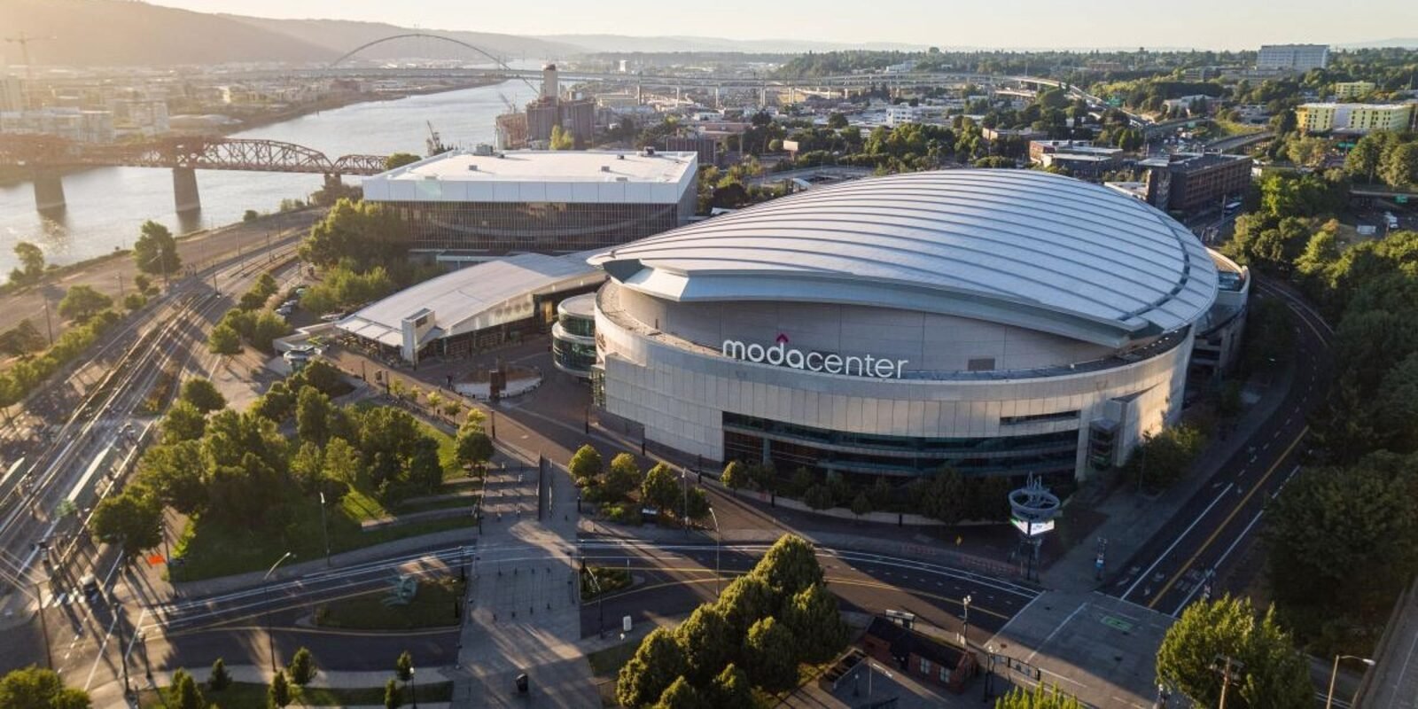 Aerial view of the Moda Center, a large modern arena with a curved roof, surrounded by roads, trees, and city buildings, near a river with bridges in the background.