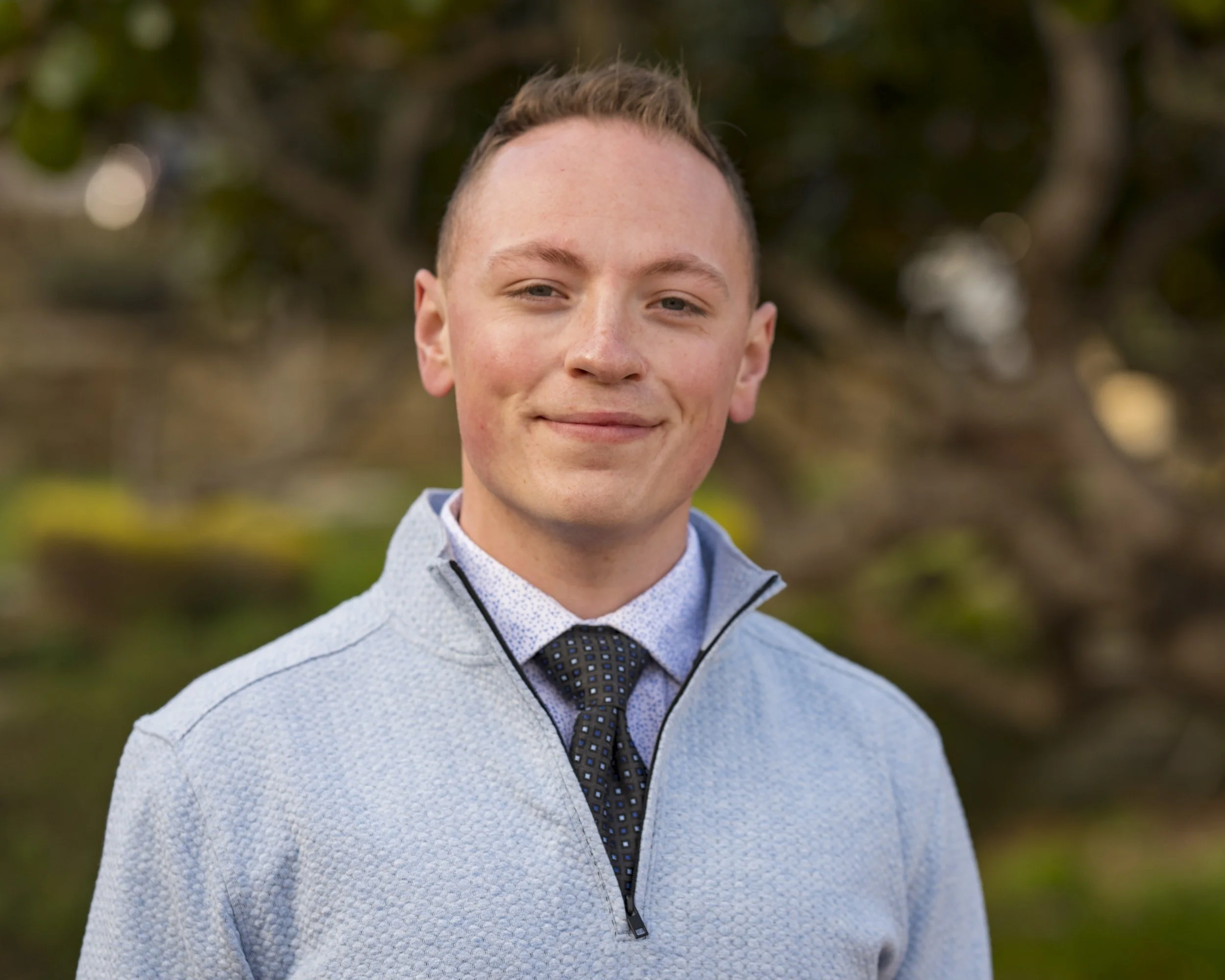 A young man outdoors with a blurred background of trees, wearing a light gray quarter-zip sweater, a collared shirt, and a patterned tie, smiling slightly.
