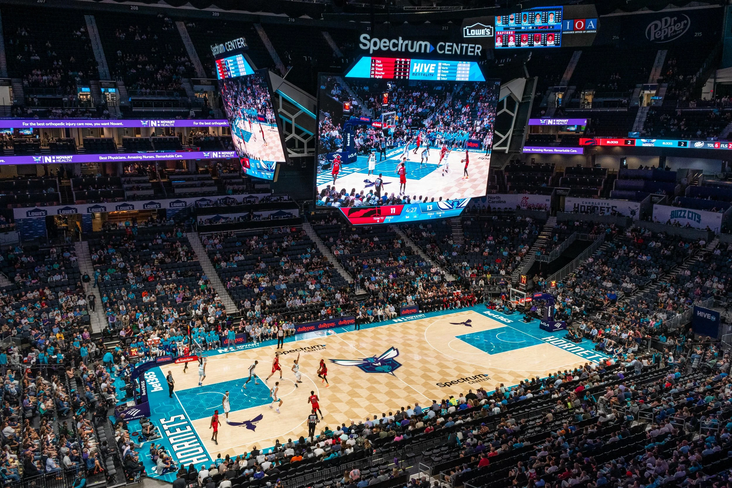 An indoor basketball game at Spectrum Center with players on the court, fans in the stands, and a large digital scoreboard displaying live game stats and video feeds.