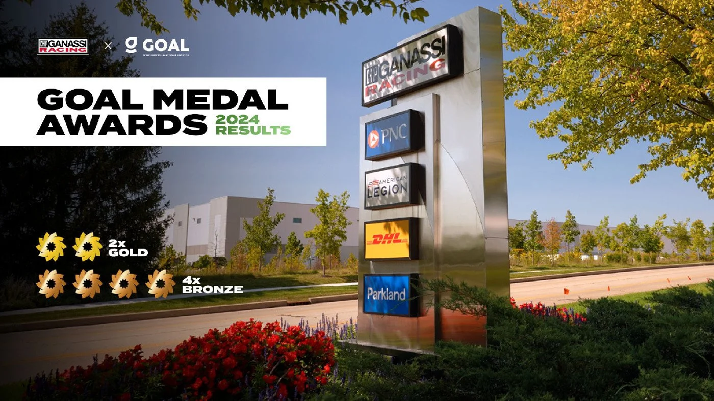 Signboard displaying 2024 Goal Medal Awards results with logos of Chip Ganassi Racing, Goal, PNC, American Legion, DHL, and Parkland, outdoors on a clear day with trees and landscaping.