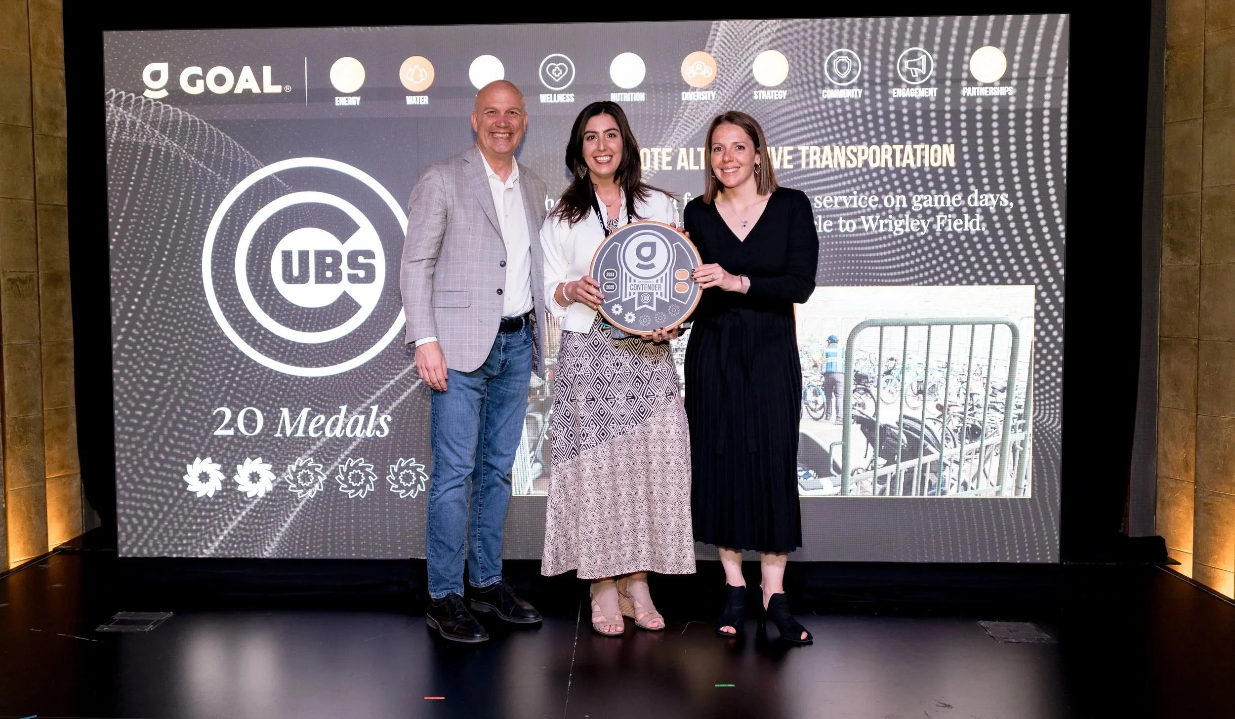 Three people standing on a stage holding a large award plaque, smiling at the camera, with a digital screen behind them displaying the GOAL logo, 20 medals, and bicycle parking options.