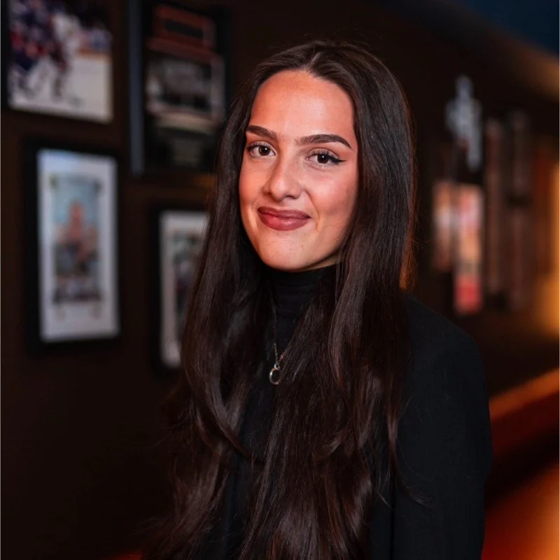 A young woman with long dark hair and a black turtleneck, smiling in a casual indoor setting with framed photos on the wall.