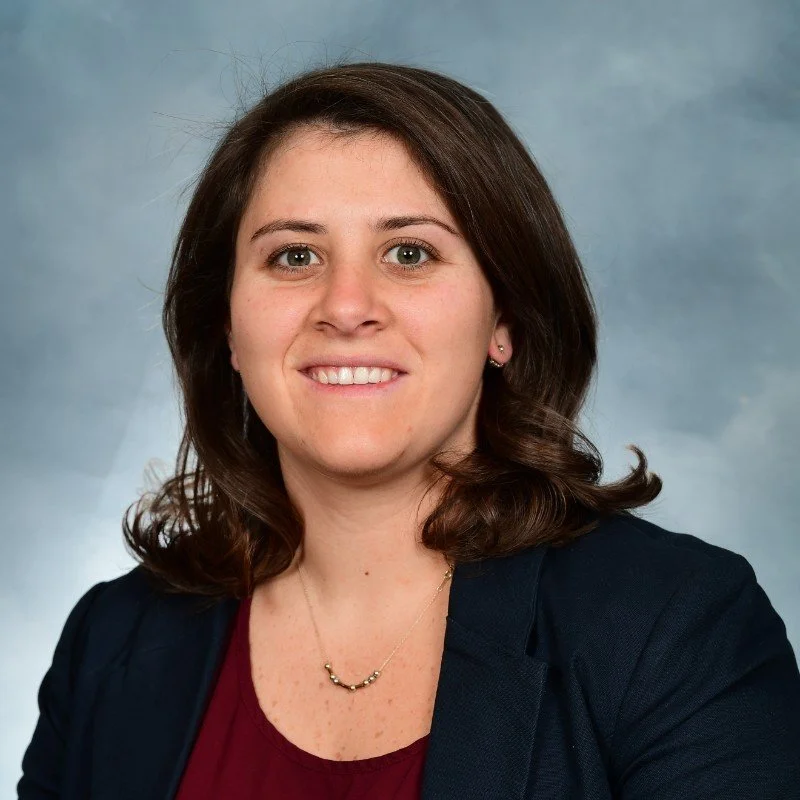 Professional woman with brown hair and green eyes smiling, wearing a navy blazer and a burgundy top, with a delicate gold necklace, against a blue-gray studio background.