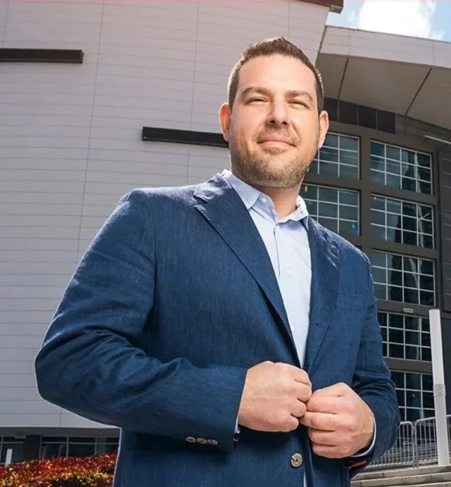 A man in a blue suit and white shirt standing outdoors in front of a modern building, facing the camera with a slight smile.