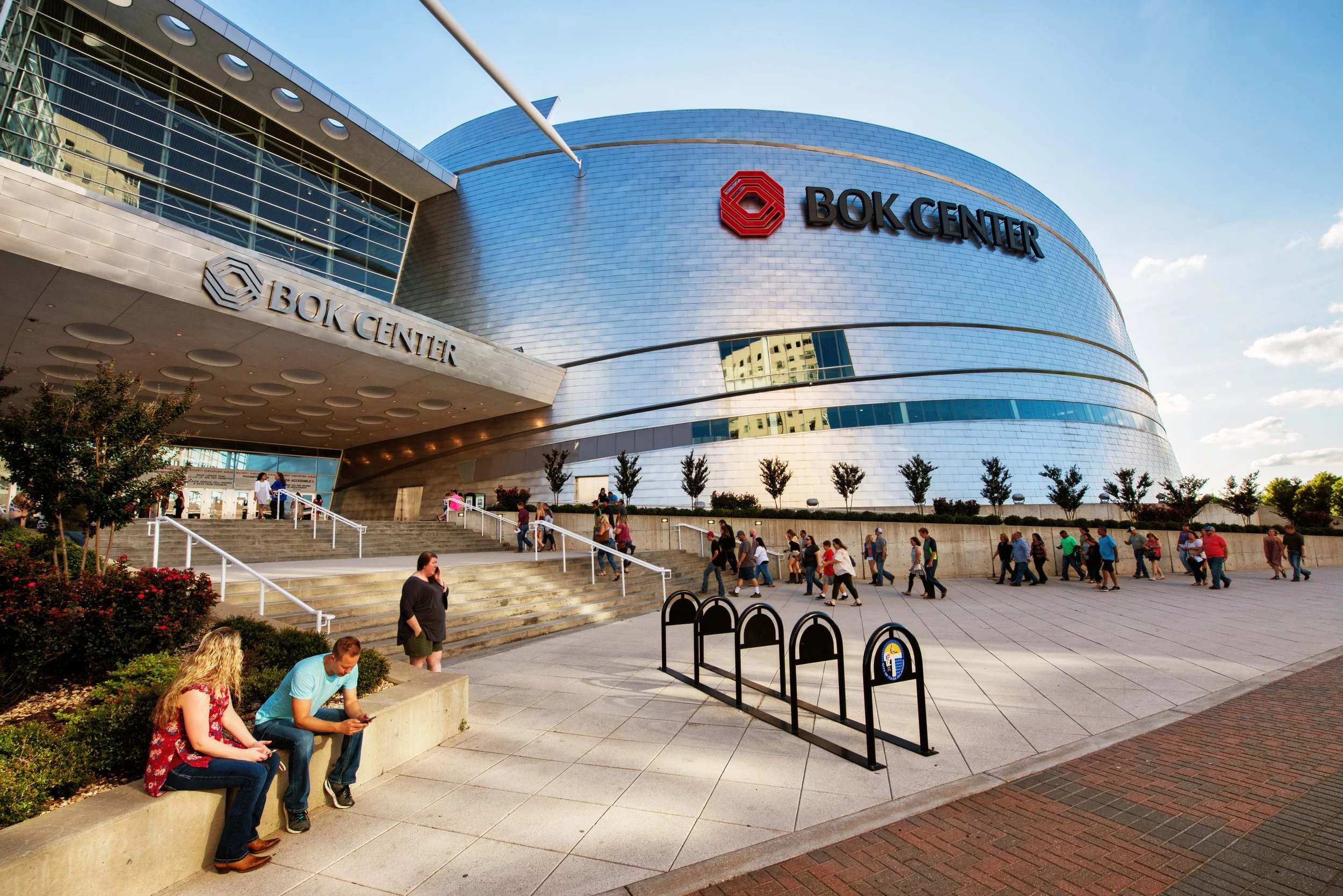 Exterior of the BOK Center, a large modern building with a shiny metallic facade. People are walking in line outside, some sitting on a ledge.