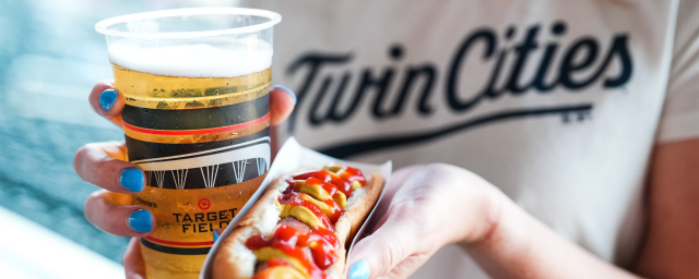 Person holding a beer in one hand and a hot dog in the other, with a blurred background.